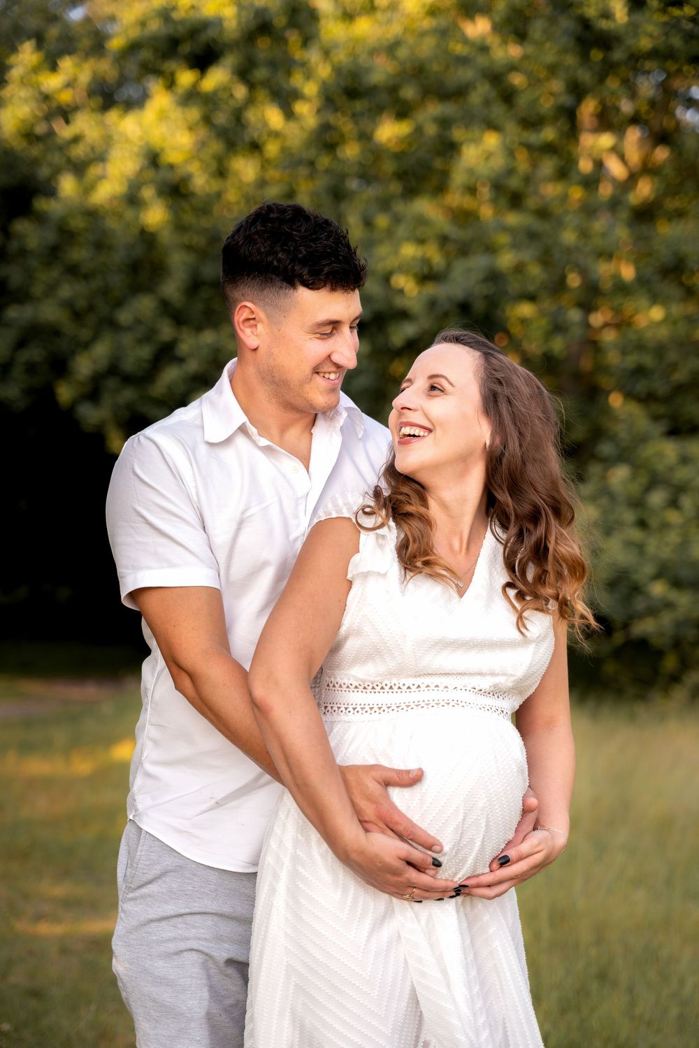 Happy pregnant couple embracing outdoors, woman in white dress cradling baby bump, man in white shirt smiling in sunny park.