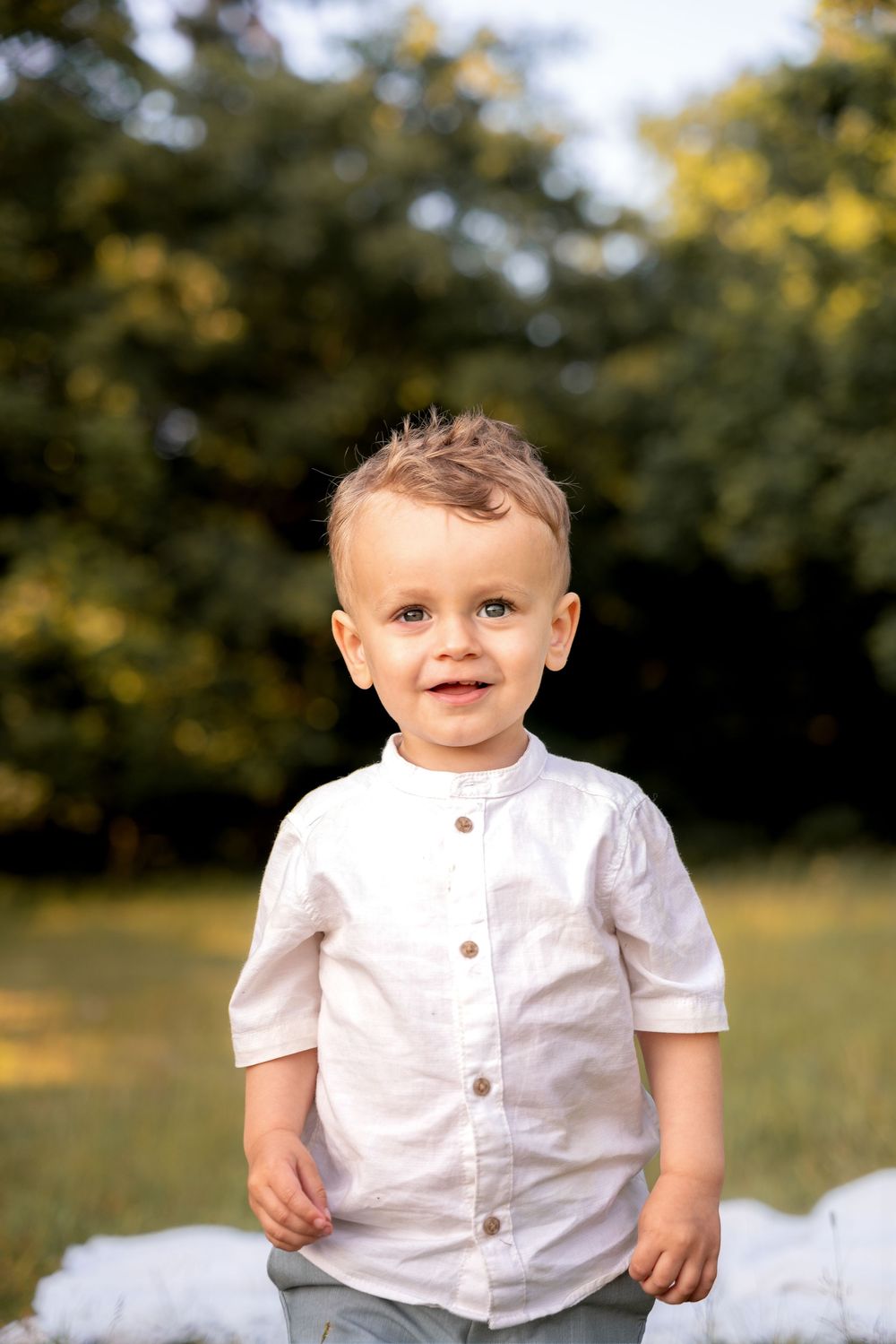 Adorable toddler boy in white linen shirt smiling outdoors in green park setting on sunny day.