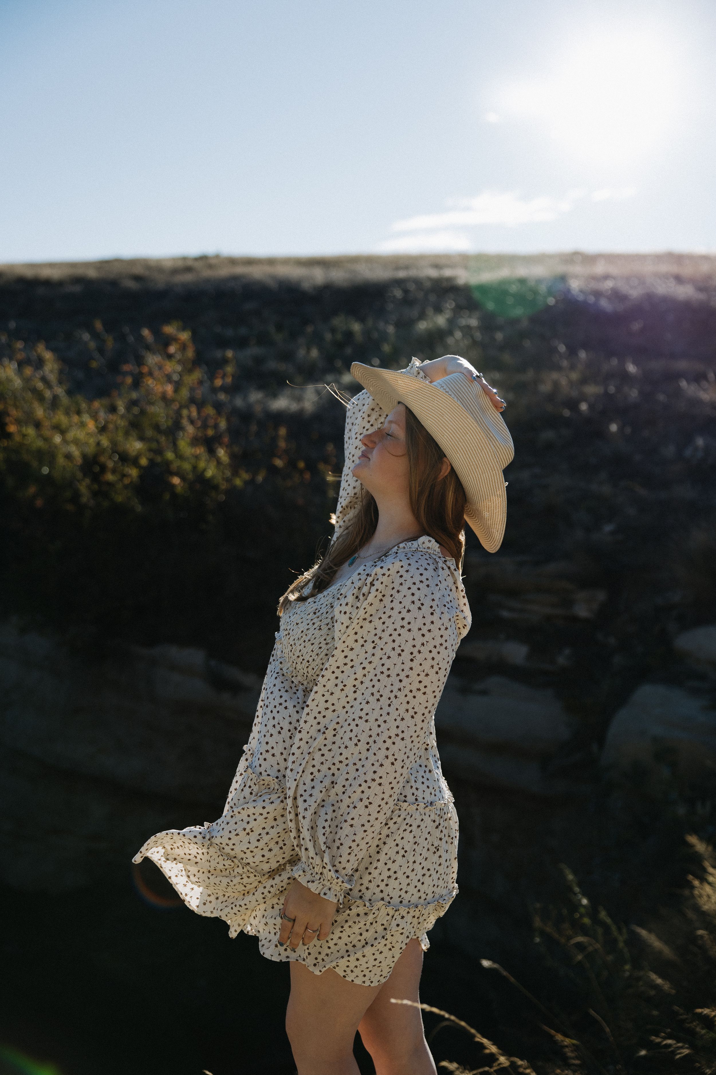 Woman in polka dot dress and cowboy hat standing in sunlight near rocky canyon landscape.