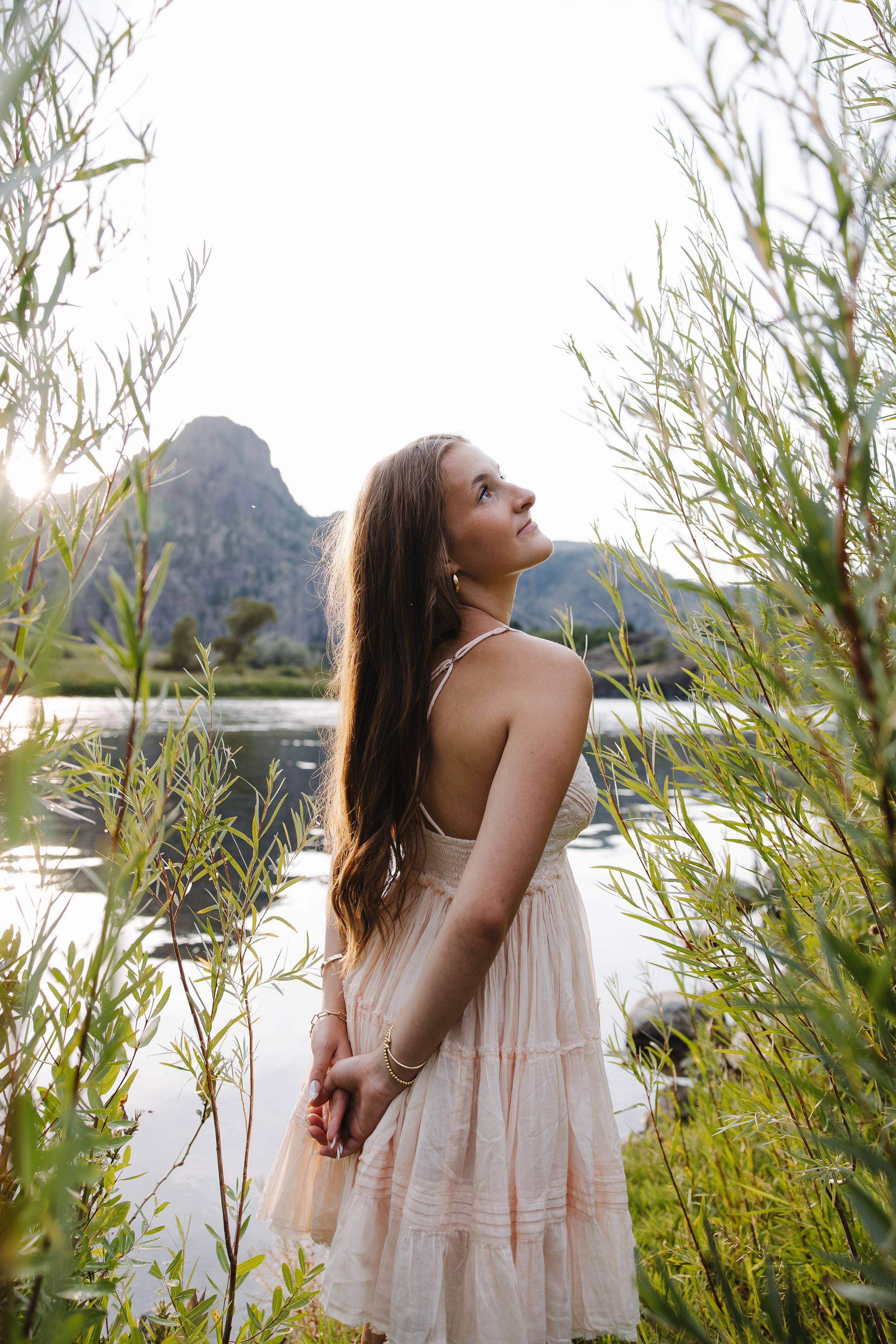 Young woman in blush spaghetti strap dress standing among tall reeds by mountain lake at sunset.