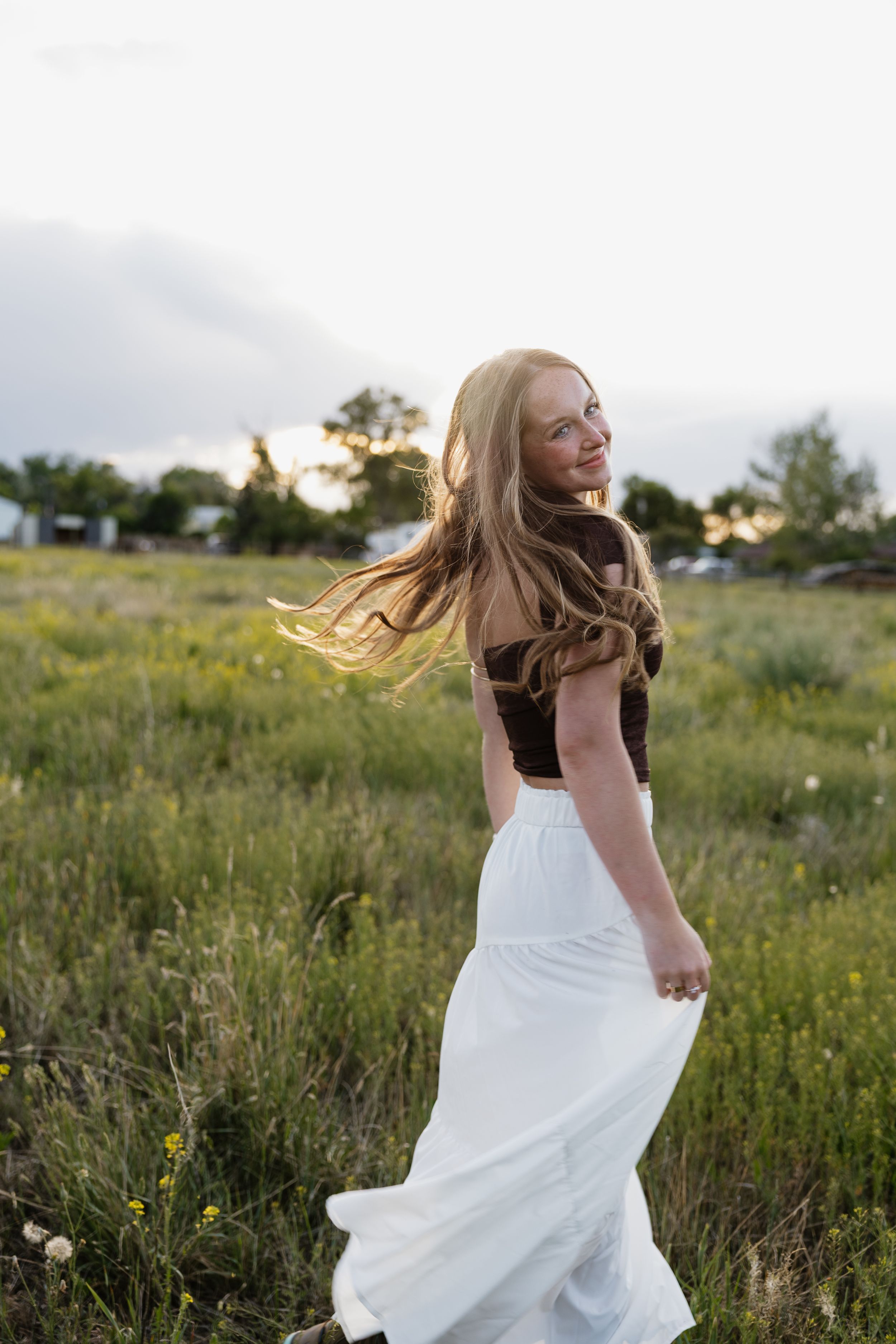 Smiling young woman in brown crop top and white skirt twirling in green meadow at sunset.