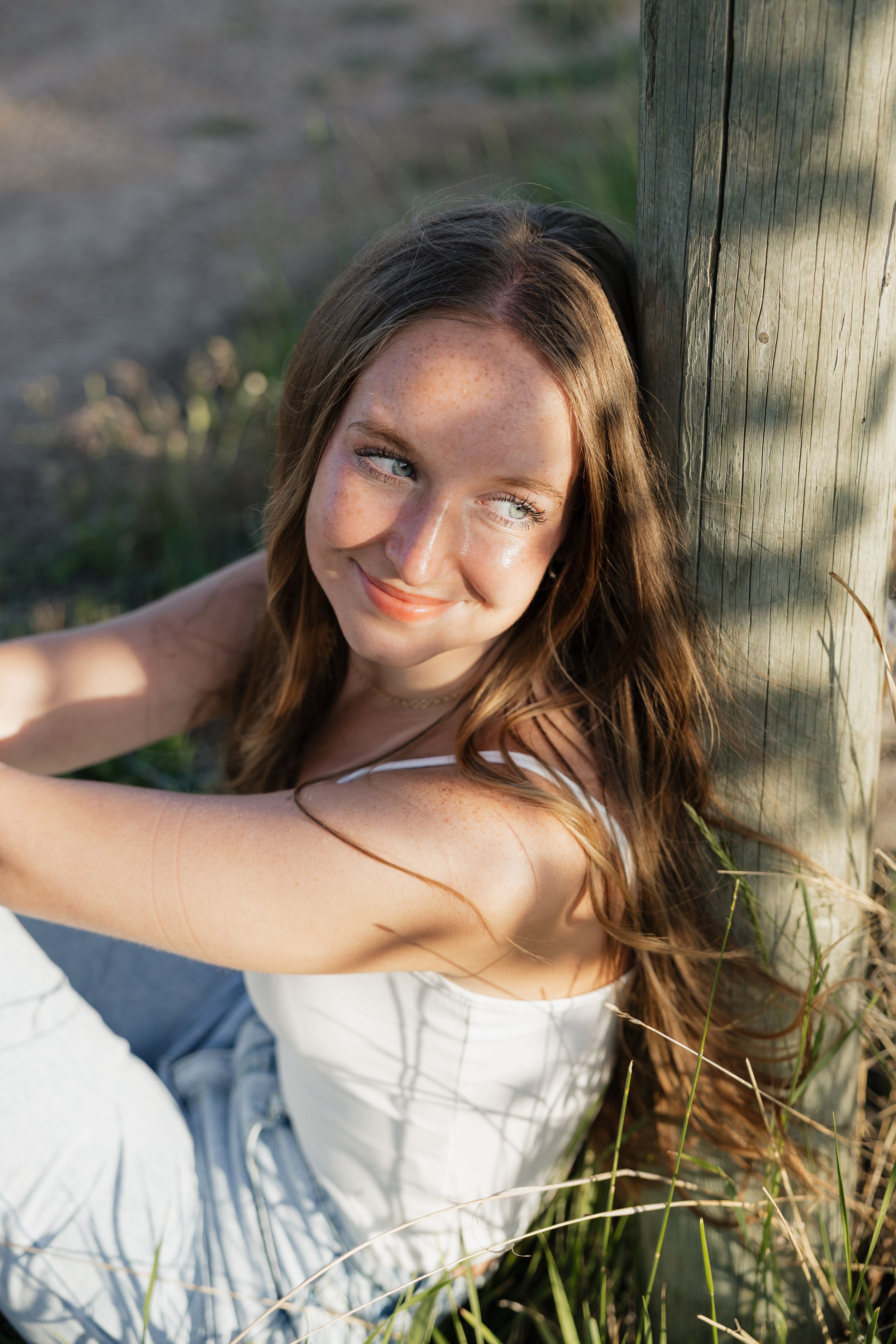 Young woman with long brown hair leaning against a wooden post smiling in warm golden sunlight.
