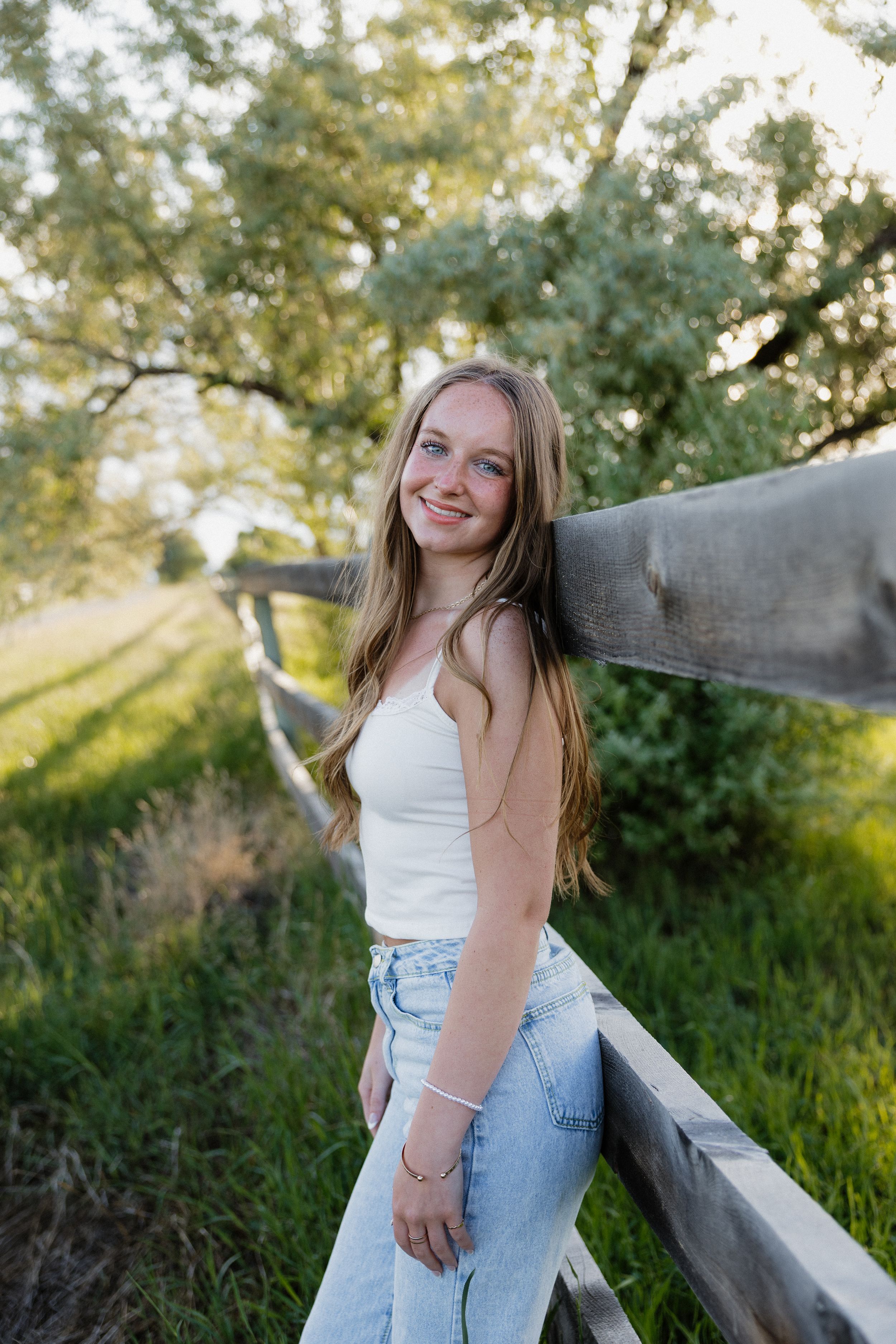 Young woman in white tank top and jeans leaning against rustic wooden fence in golden sunlight.