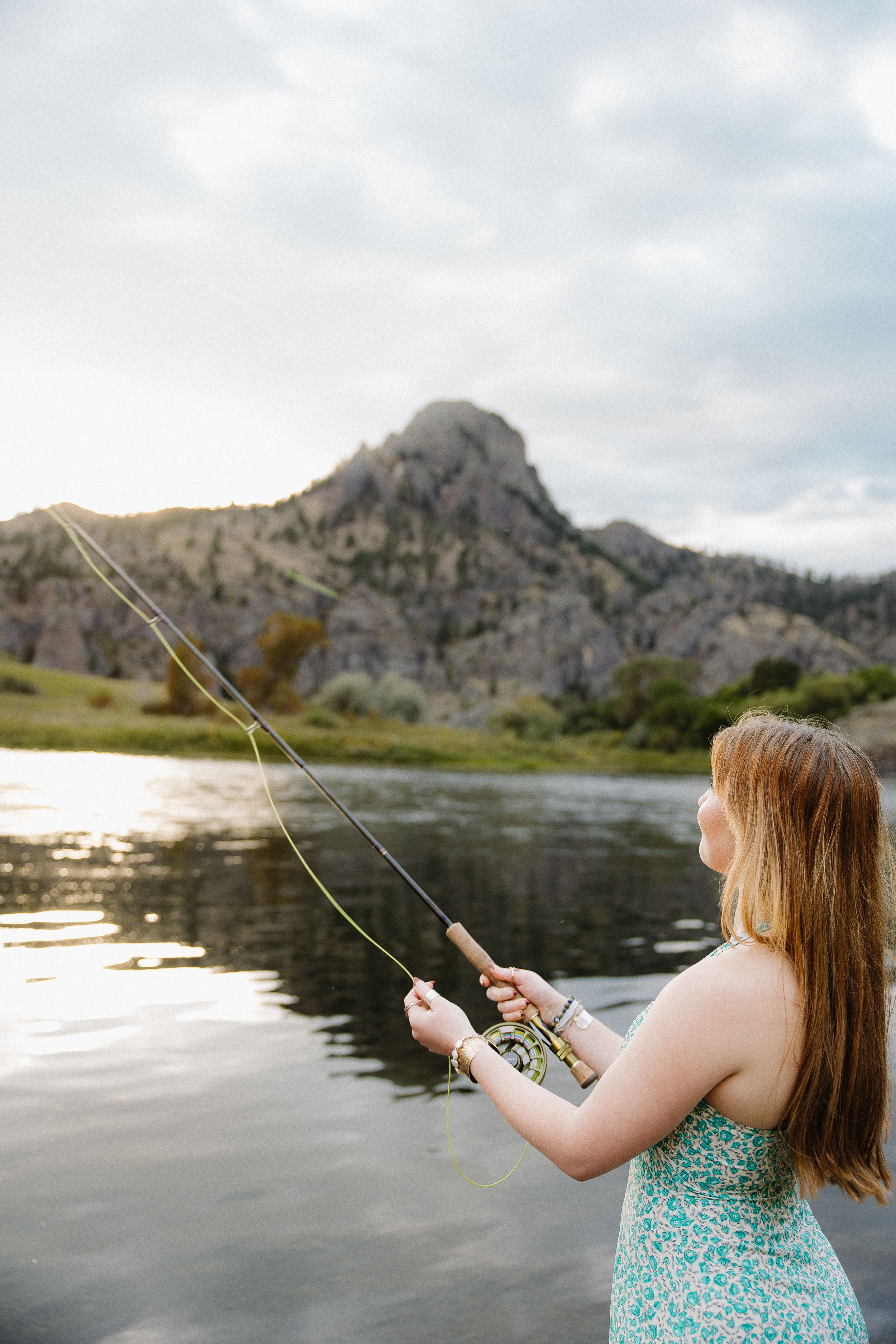 Young woman in floral dress fly fishing on calm river with mountain landscape behind her.