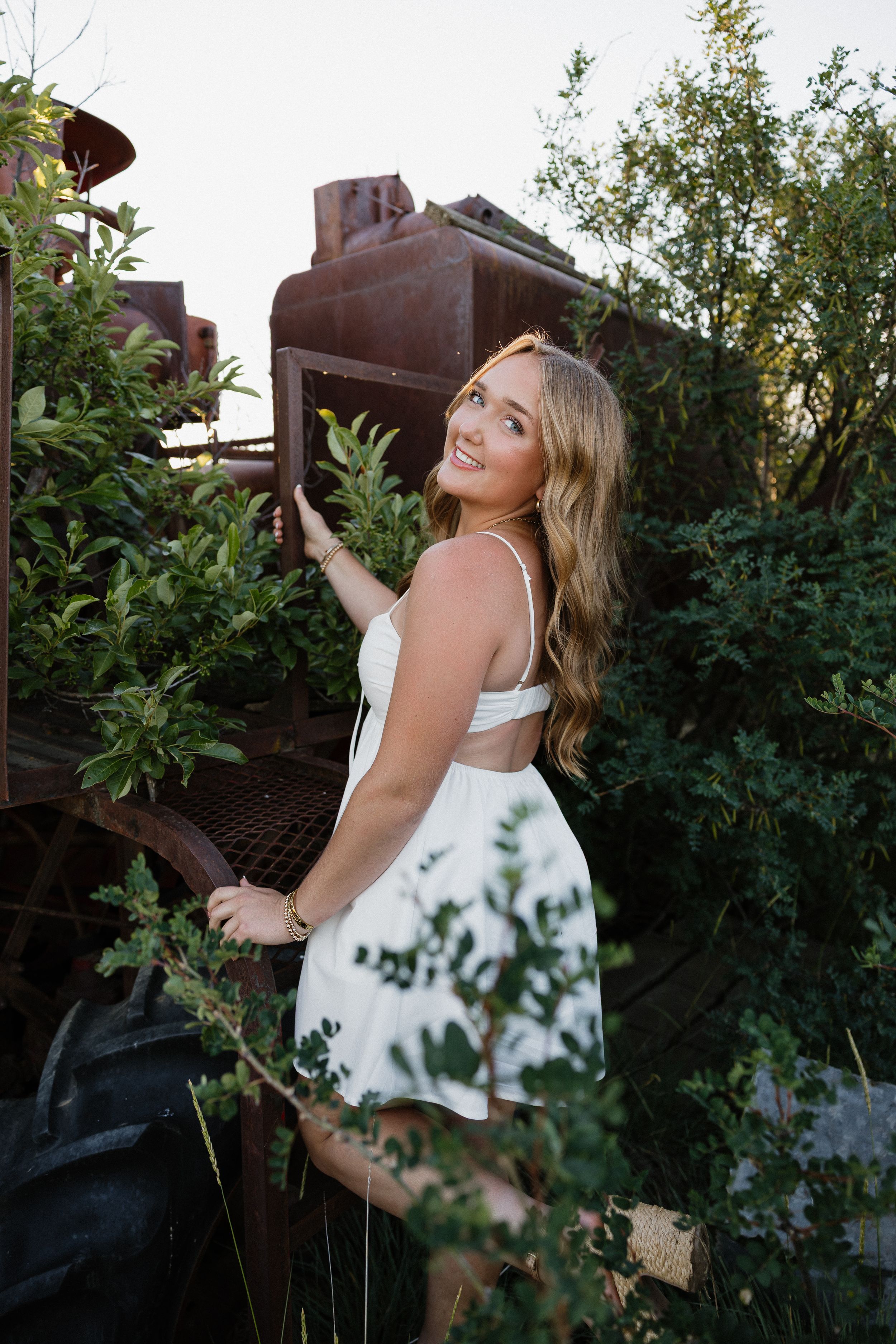Smiling young woman in white dress climbing rustic vintage farm equipment surrounded by green foliage.