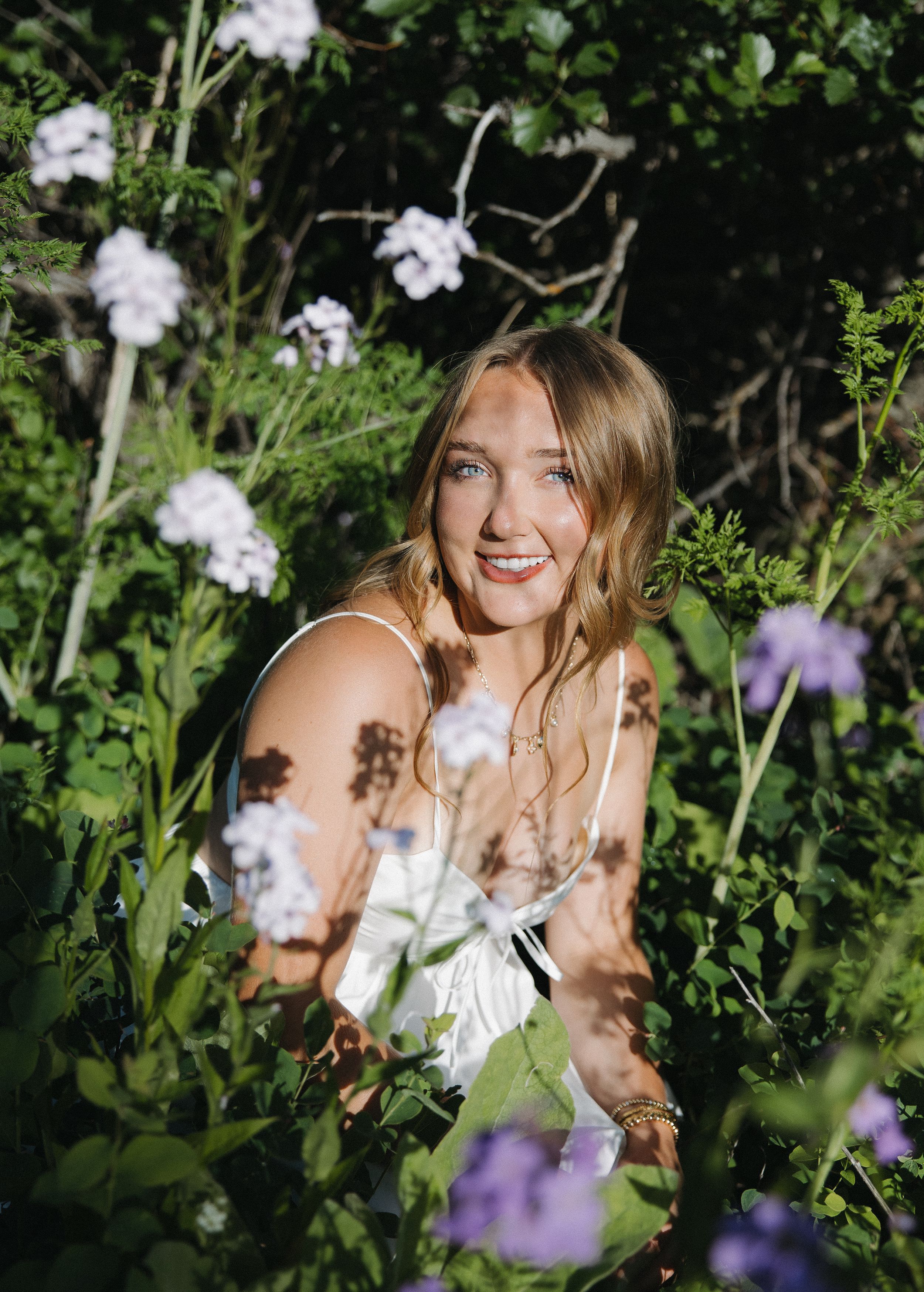 Young blonde woman smiling among white and purple wildflowers in dappled sunlight.