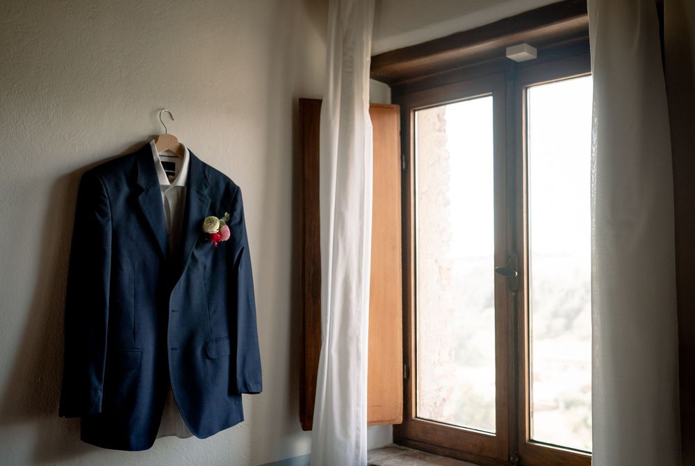 Navy blue groom suit jacket with floral boutonniere hanging on a window frame in a rustic room.
