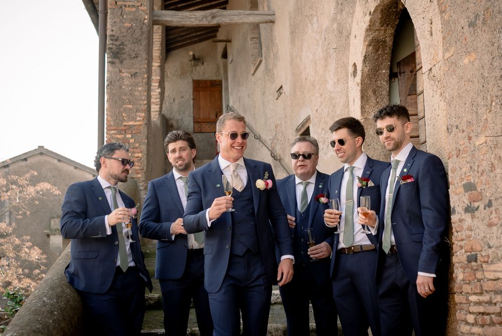 Groom and groomsmen in navy suits celebrating with champagne outside a rustic stone wedding venue.