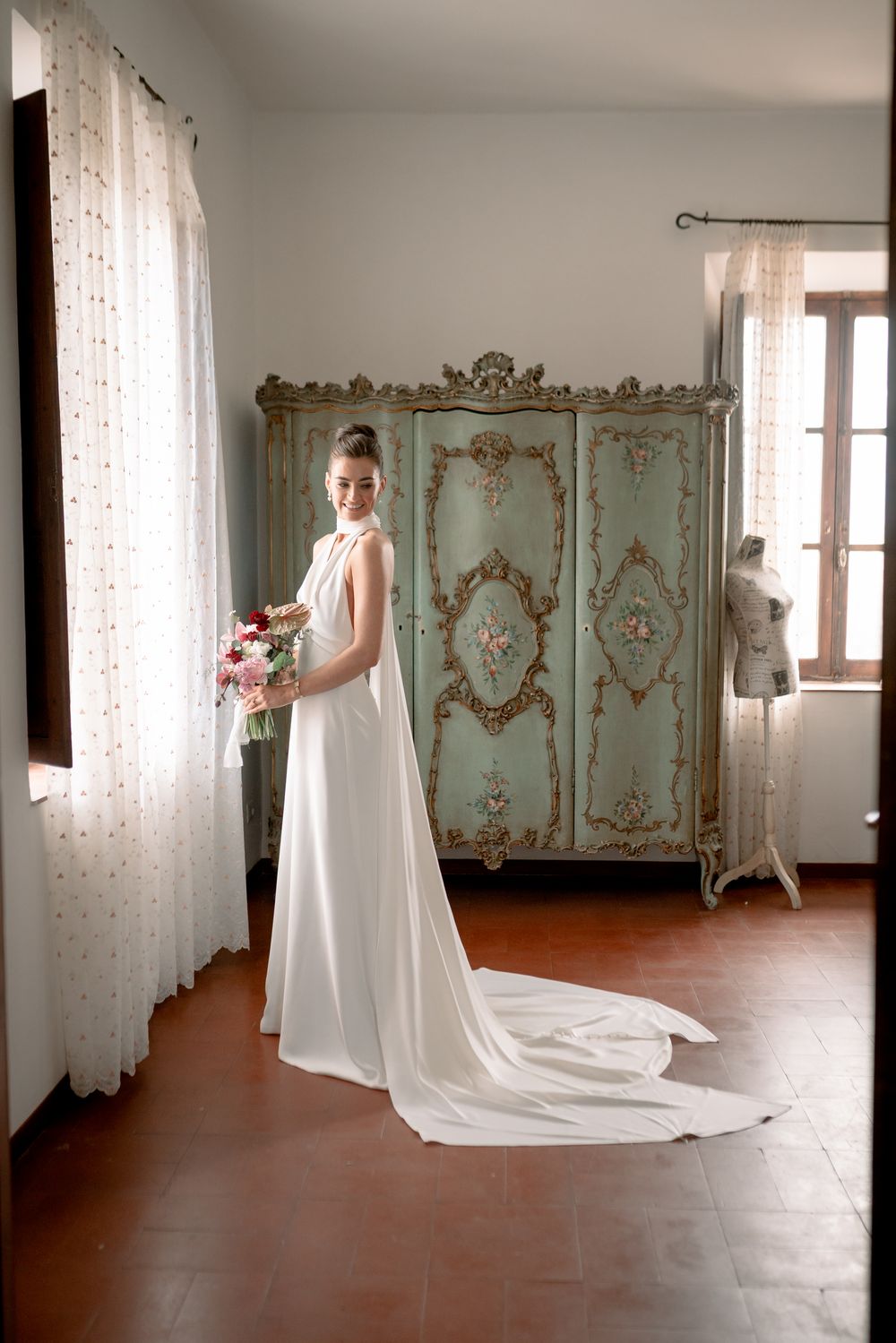Bride in elegant white halter gown with long train holds bouquet beside ornate vintage armoire in sunlit room.