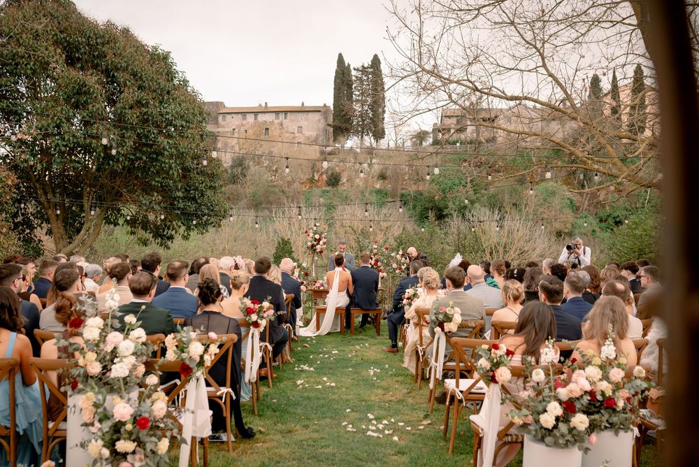 Outdoor wedding ceremony at Tuscan estate with guests seated along flower-lined aisle amid lush gardens.