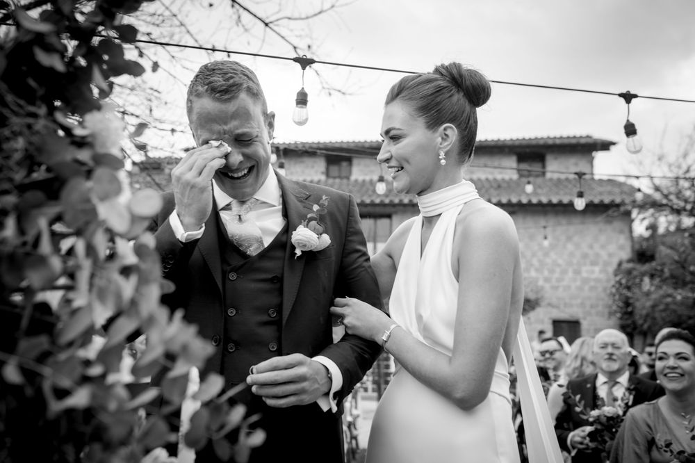 Black and white photo of groom wiping tears while bride smiles during joyful wedding ceremony moment.