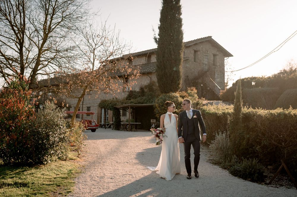 Newlyweds walk hand in hand along garden path with rustic Tuscan farmhouse in golden hour light.