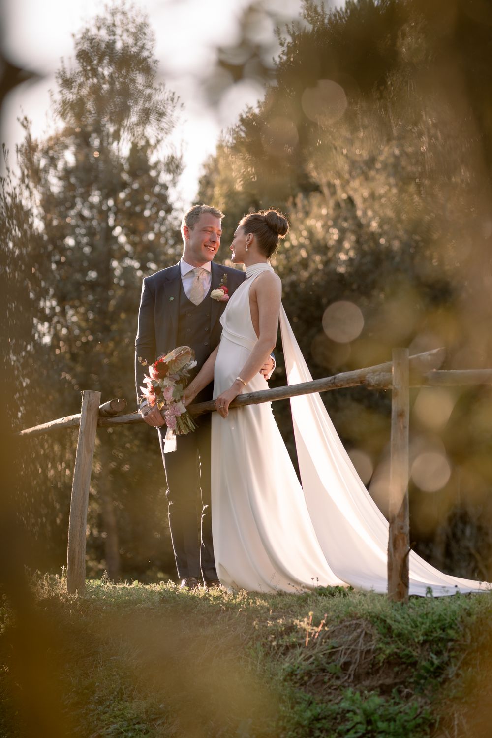 Smiling bride and groom pose by a rustic wooden fence surrounded by golden olive trees at sunset.