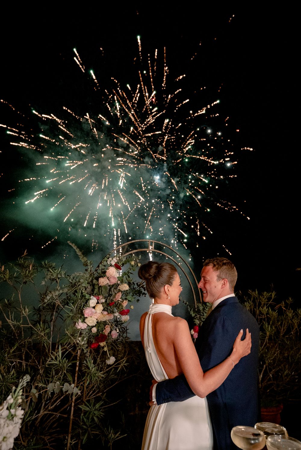 Bride and groom embrace romantically as colorful fireworks explode above a floral arch at night.