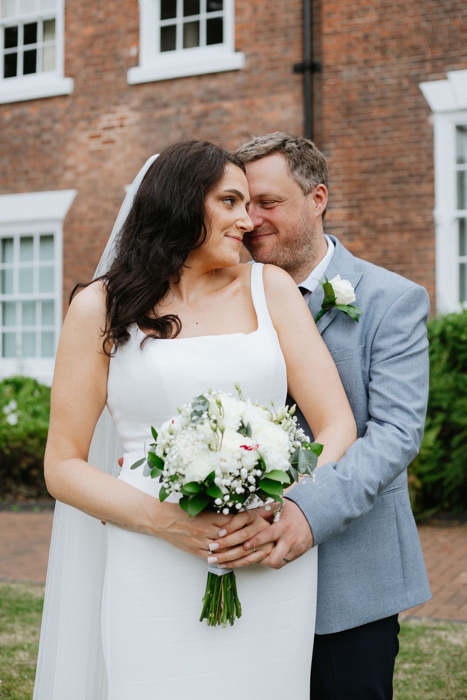 Bride in white gown holding bouquet embraced by groom in gray suit outside brick building.