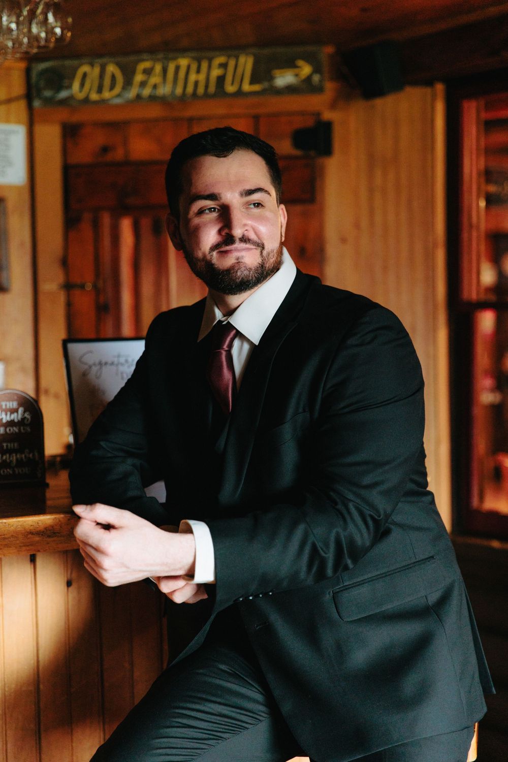 Handsome groom in black suit with burgundy tie smiling at camera inside rustic wooden venue with Old Faithful sign.