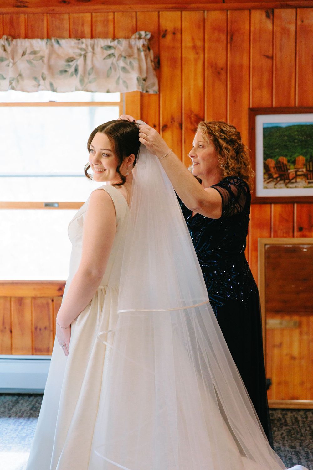Mother helps bride with veil in rustic wood-paneled room before wedding ceremony.