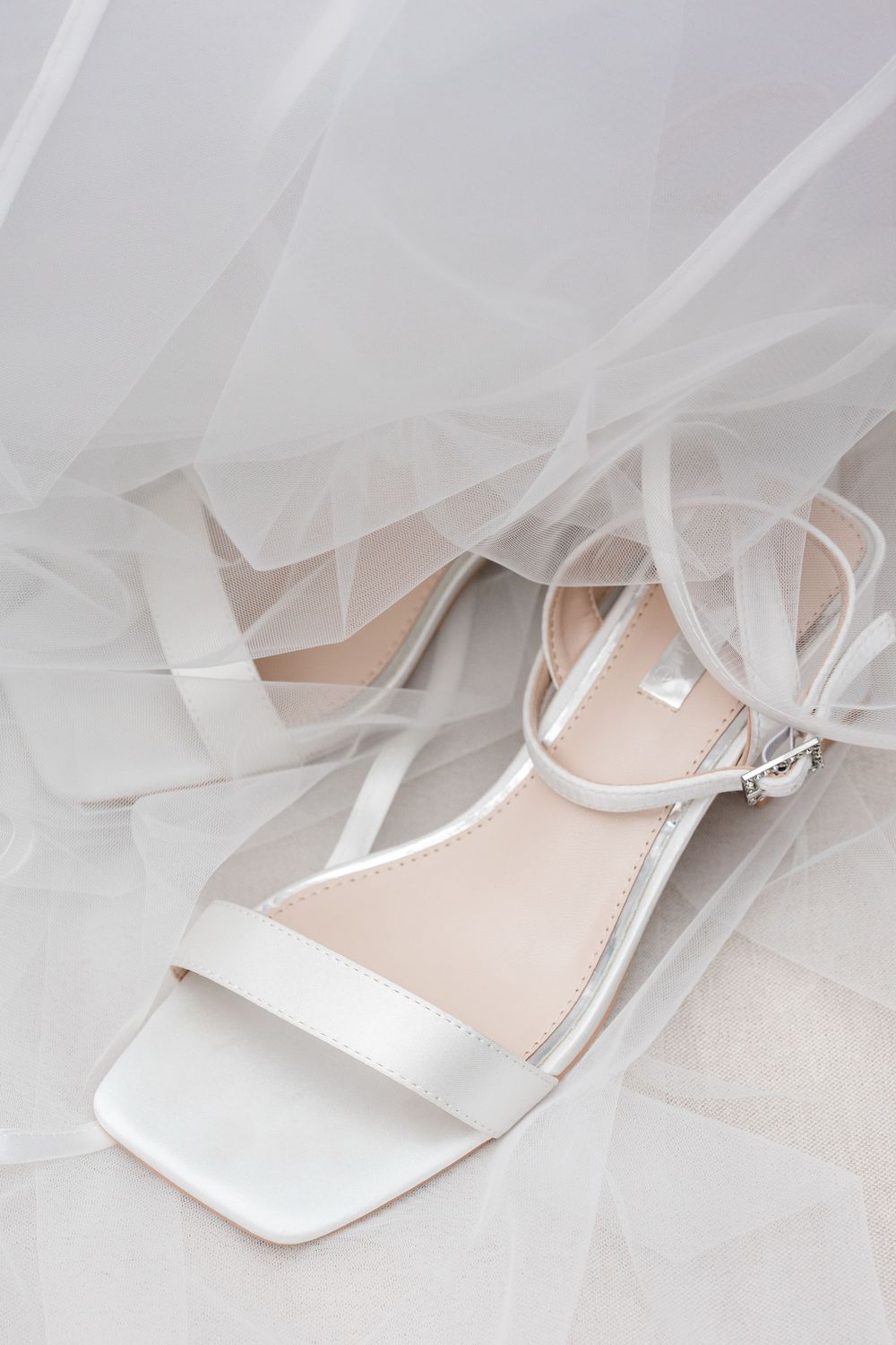 White satin bridal heeled sandals with ankle strap draped in sheer wedding veil on light background.