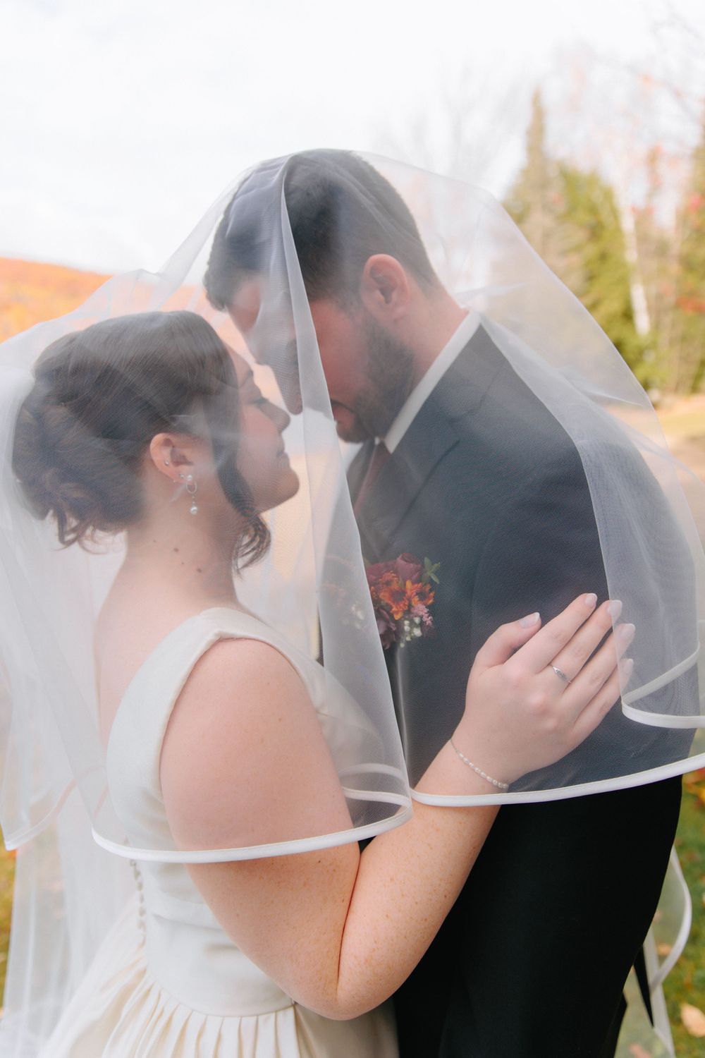 Bride and groom sharing intimate moment under sheer wedding veil surrounded by autumn foliage outdoors.