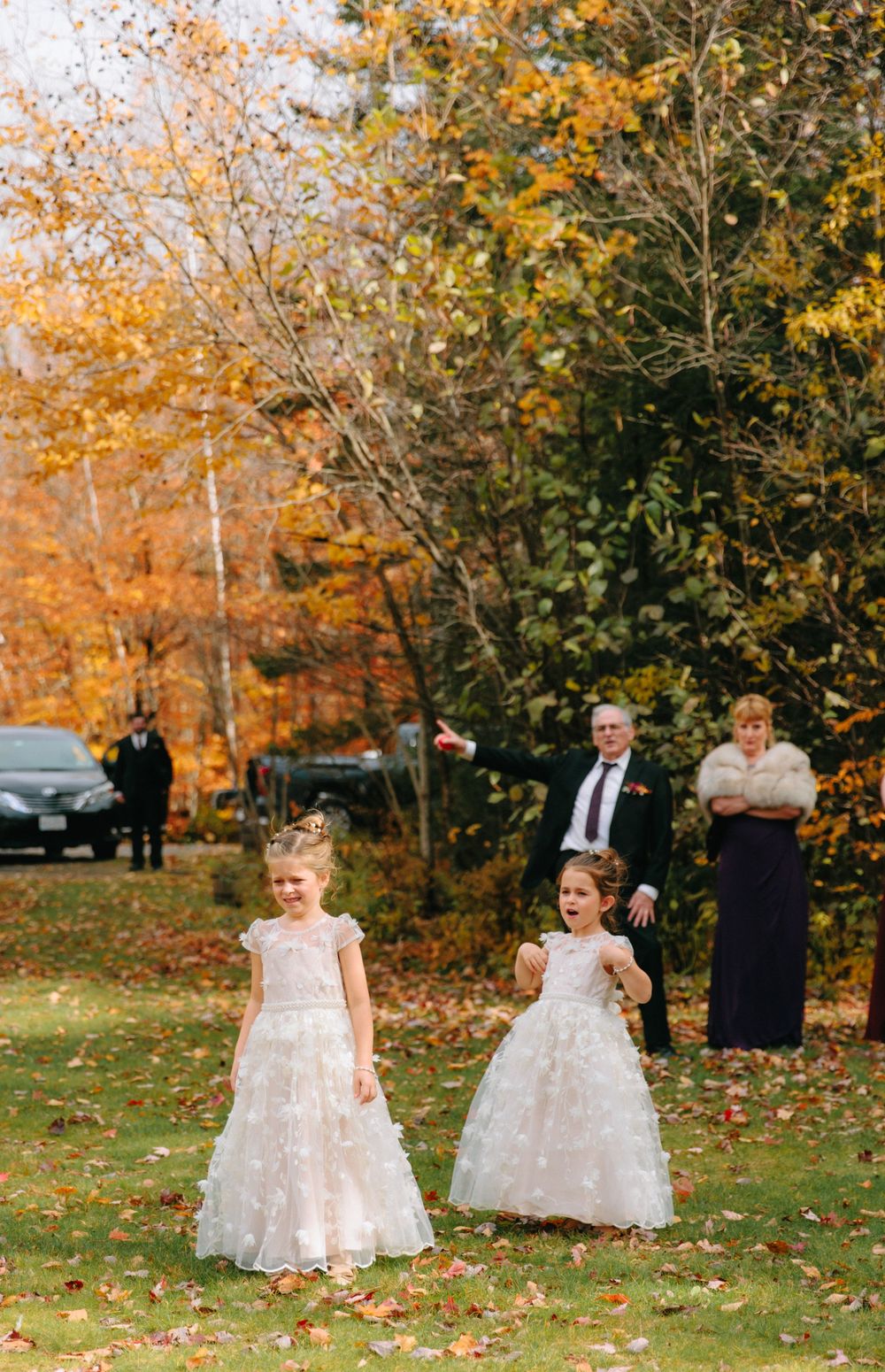 Two flower girls in white lace dresses tossing petals on grass amid golden autumn trees at outdoor wedding.