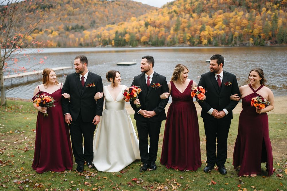 Bridal party in burgundy dresses and black suits walk by an autumn lake, bride holding orange bouquet.