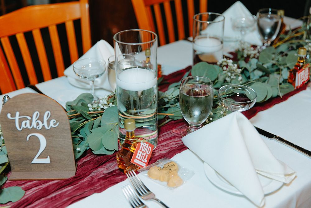 Elegant wedding table setting with eucalyptus garland, burgundy runner, white napkins, and a wooden Table 2 sign.