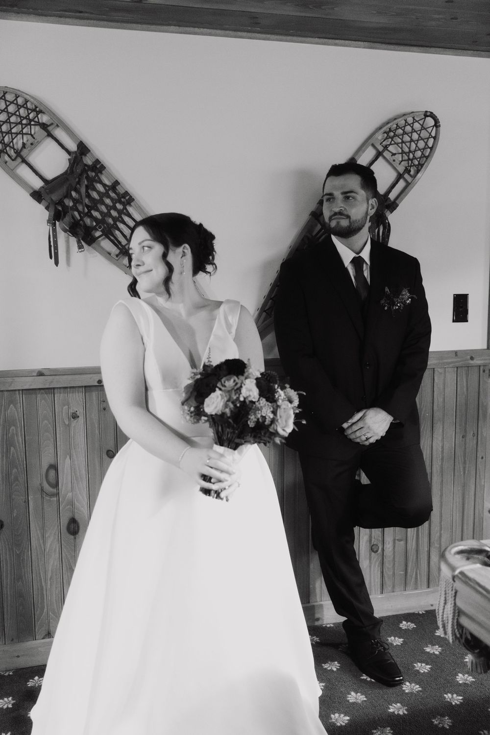 Bride holding bouquet and groom lean against wall with snowshoes in black and white wedding photo.