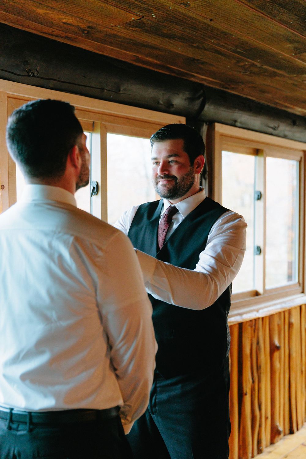 Groom smiles while groomsman adjusts his vest inside rustic wooden cabin before wedding ceremony.