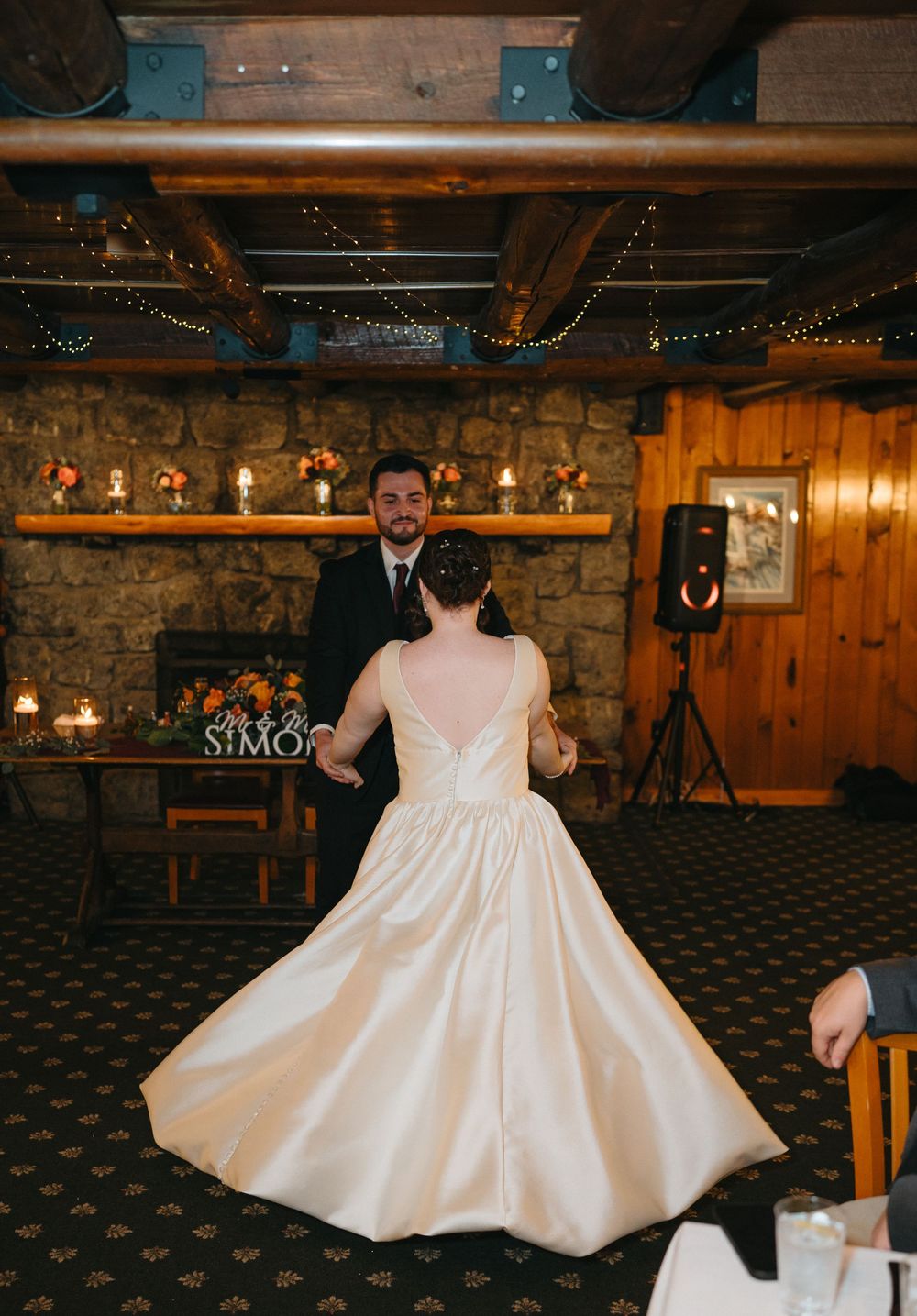 Bride twirls in elegant white gown during first dance near stone fireplace in rustic reception venue.