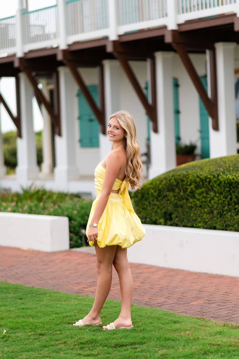 Young blonde woman in yellow sundress smiling outdoors on green lawn near white building with teal shutters.