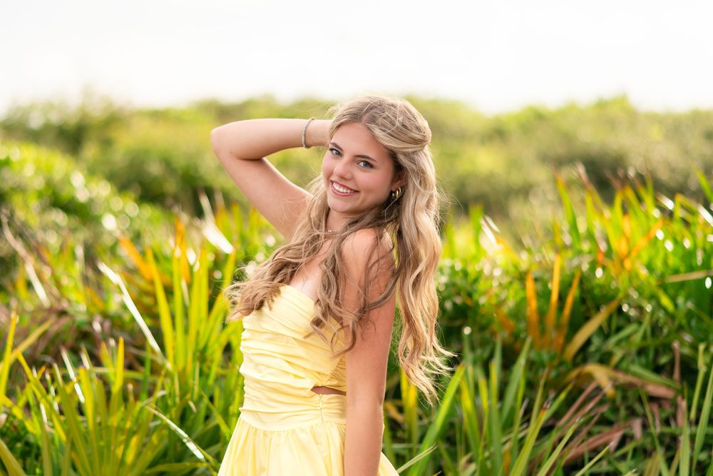 Young blonde woman in yellow dress smiling in lush green field, golden hour outdoor portrait photography.