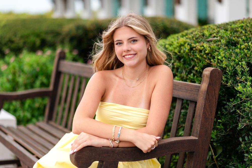 Smiling young woman in yellow strapless dress sitting on wooden bench outdoors with green hedges background.