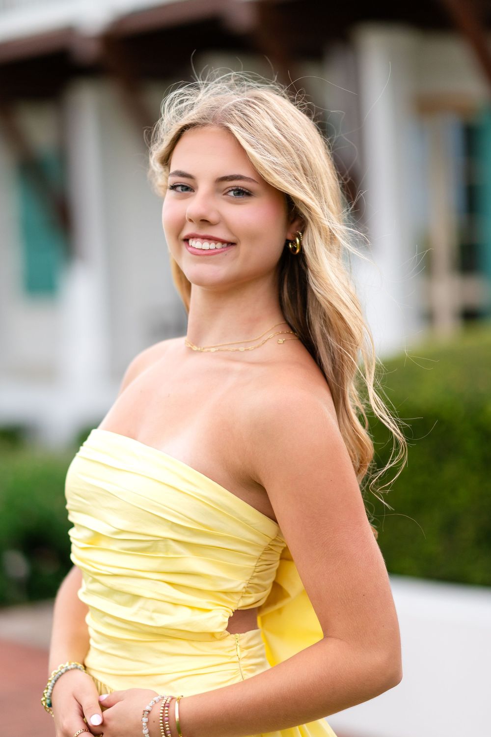 Young blonde woman smiling in a yellow strapless dress, wearing gold jewelry outdoors.