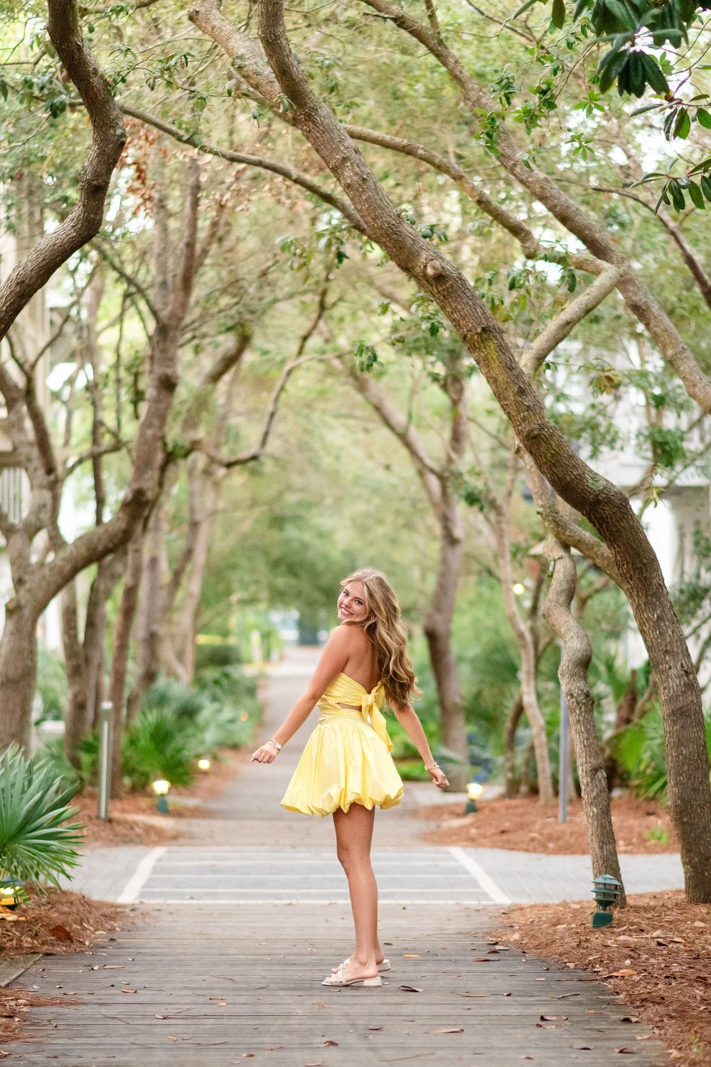 Young woman in yellow dress twirling on a tree-lined boardwalk path.
