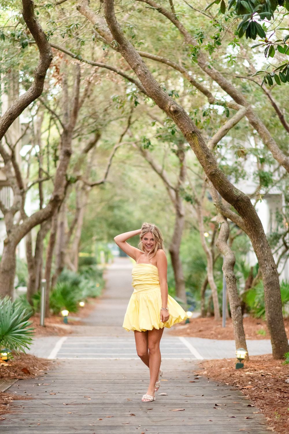 Blonde woman in yellow mini dress walking on tree-lined boardwalk path in lush green setting.