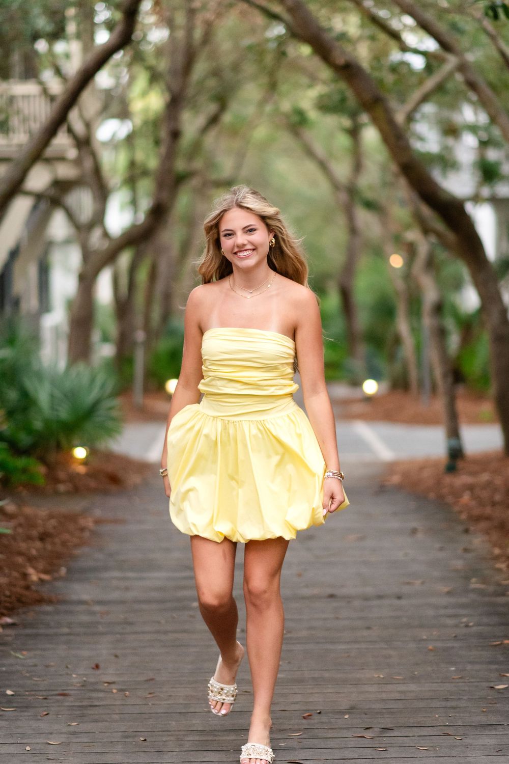 Young woman in yellow strapless bubble dress walking on a tree-lined boardwalk path, smiling confidently.