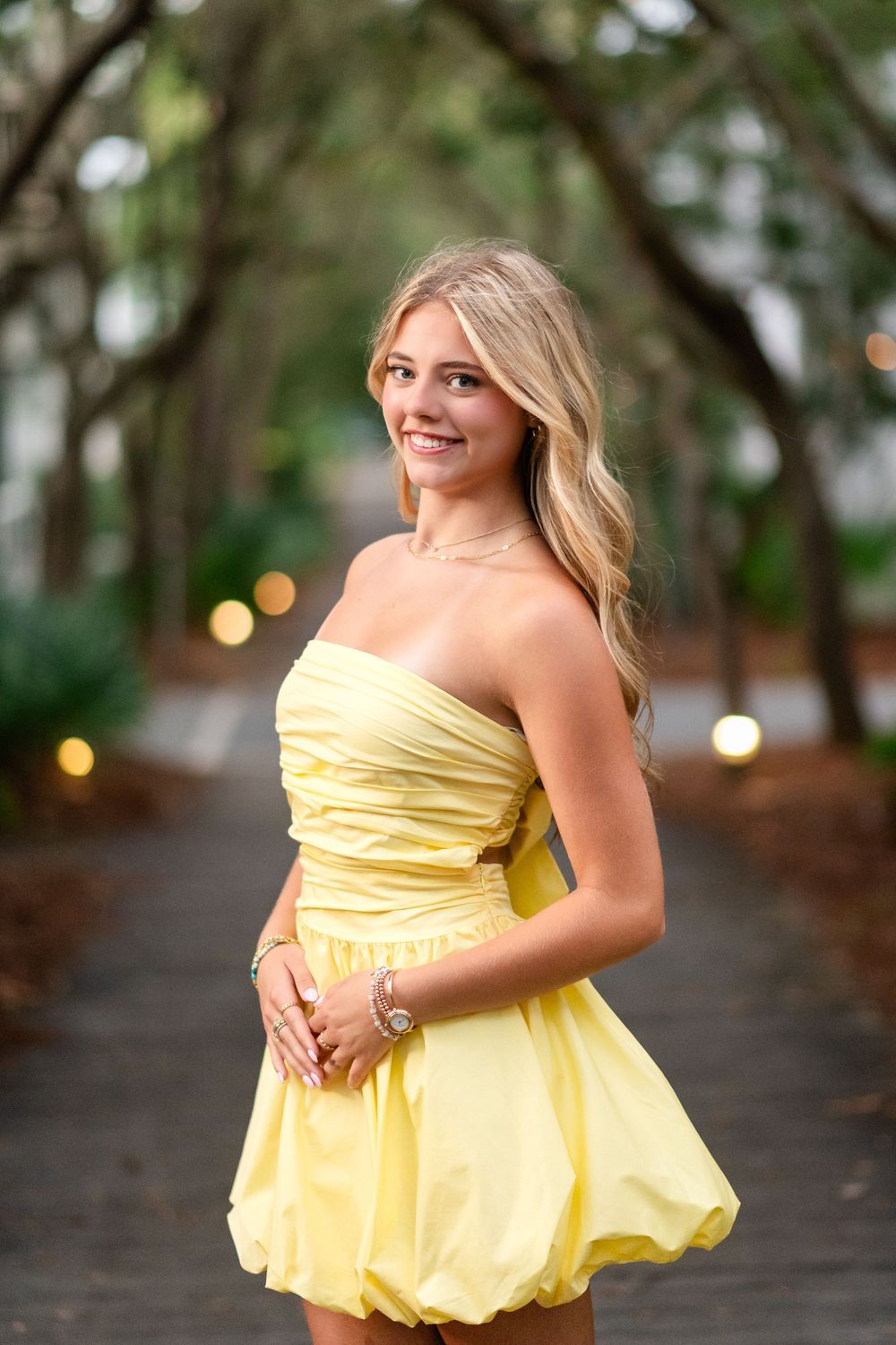 Smiling blonde woman in strapless yellow bubble dress on bokeh-lit path.