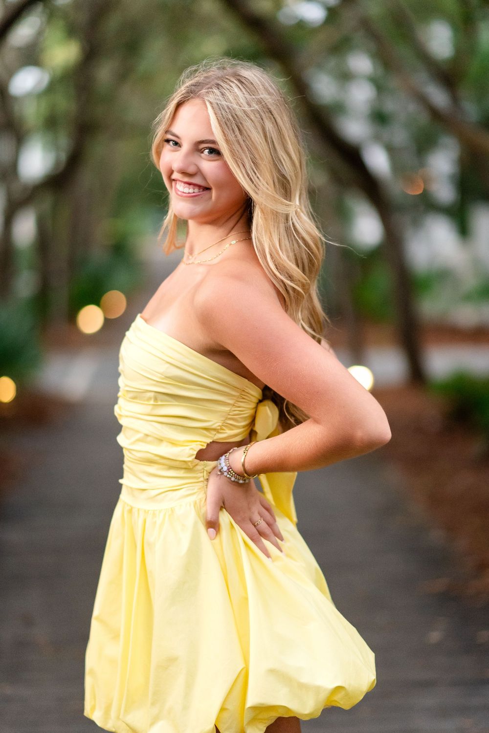 Happy blonde woman in yellow strapless dress twirling on tree-lined path with bokeh lights background.