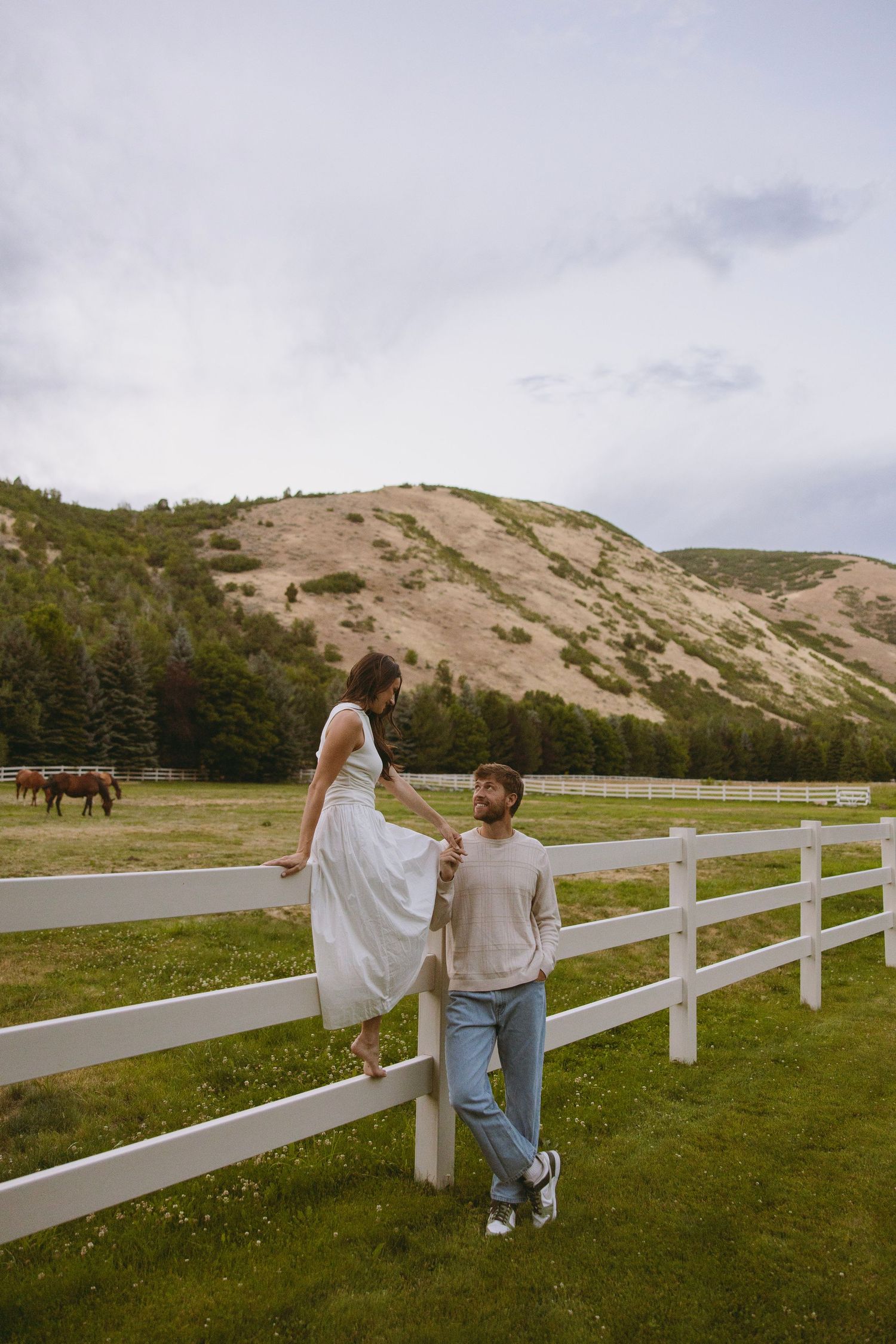 A bride in a flowing white dress stands gracefully on a wooden deck surrounded by evergreen mountains at sunset.