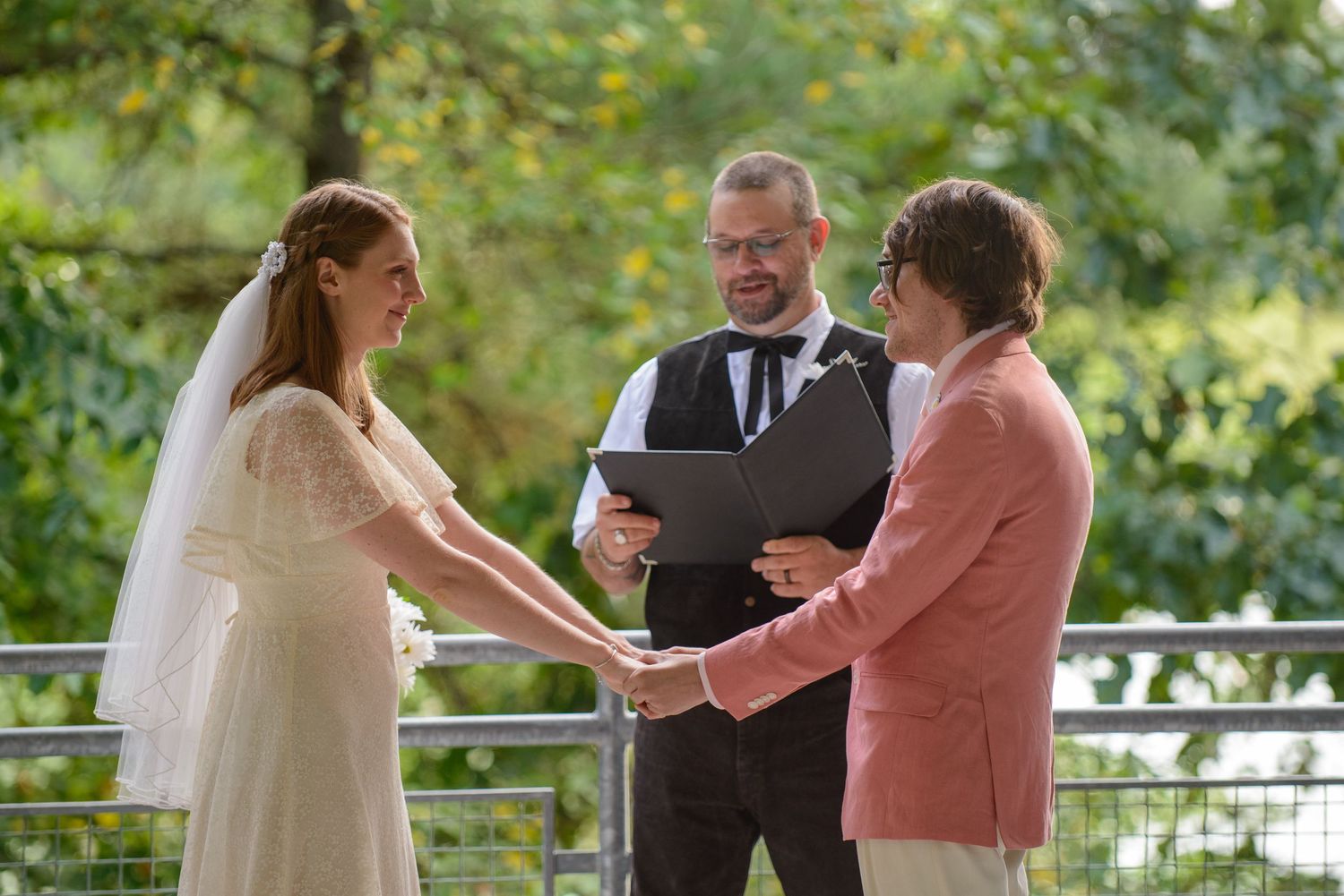 Bride and partner hold hands during outdoor wedding ceremony officiated by man in black vest.