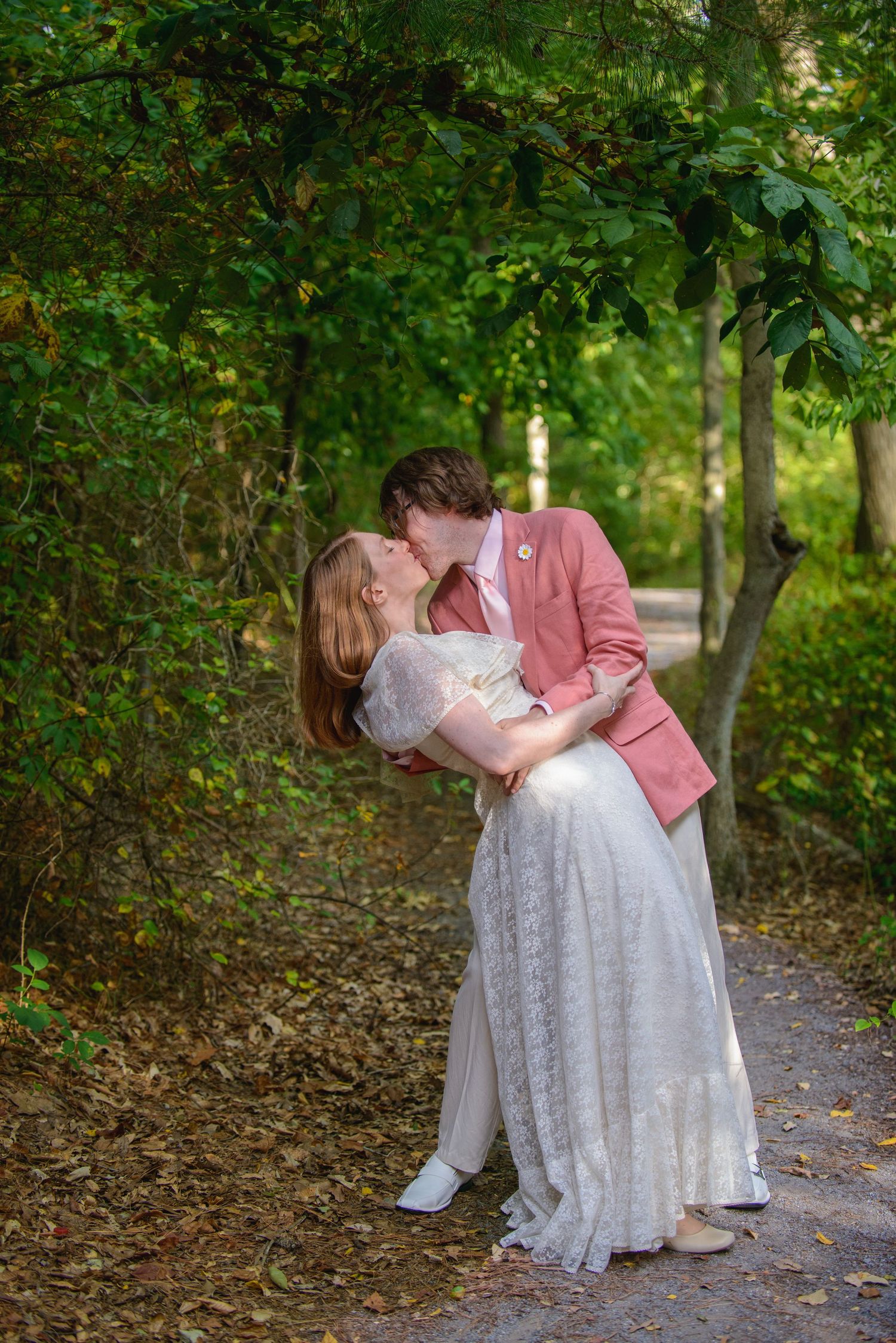Couple sharing a romantic kiss on a tree-lined path, wearing wedding attire in soft pink and white.