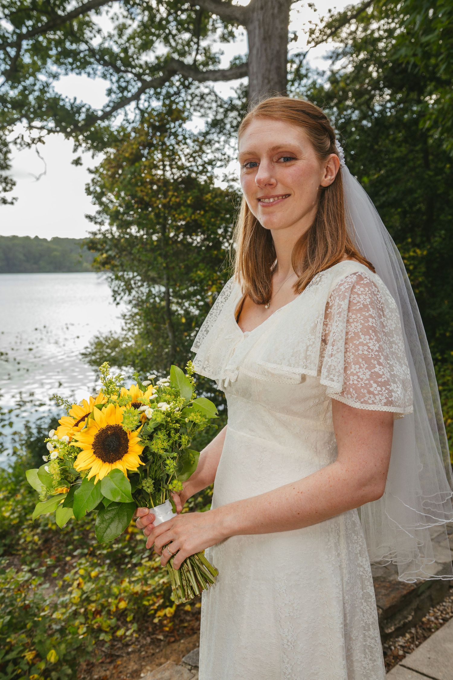 Smiling bride in white lace dress holding sunflower bouquet with veil, standing by a serene lake surrounded by trees.
