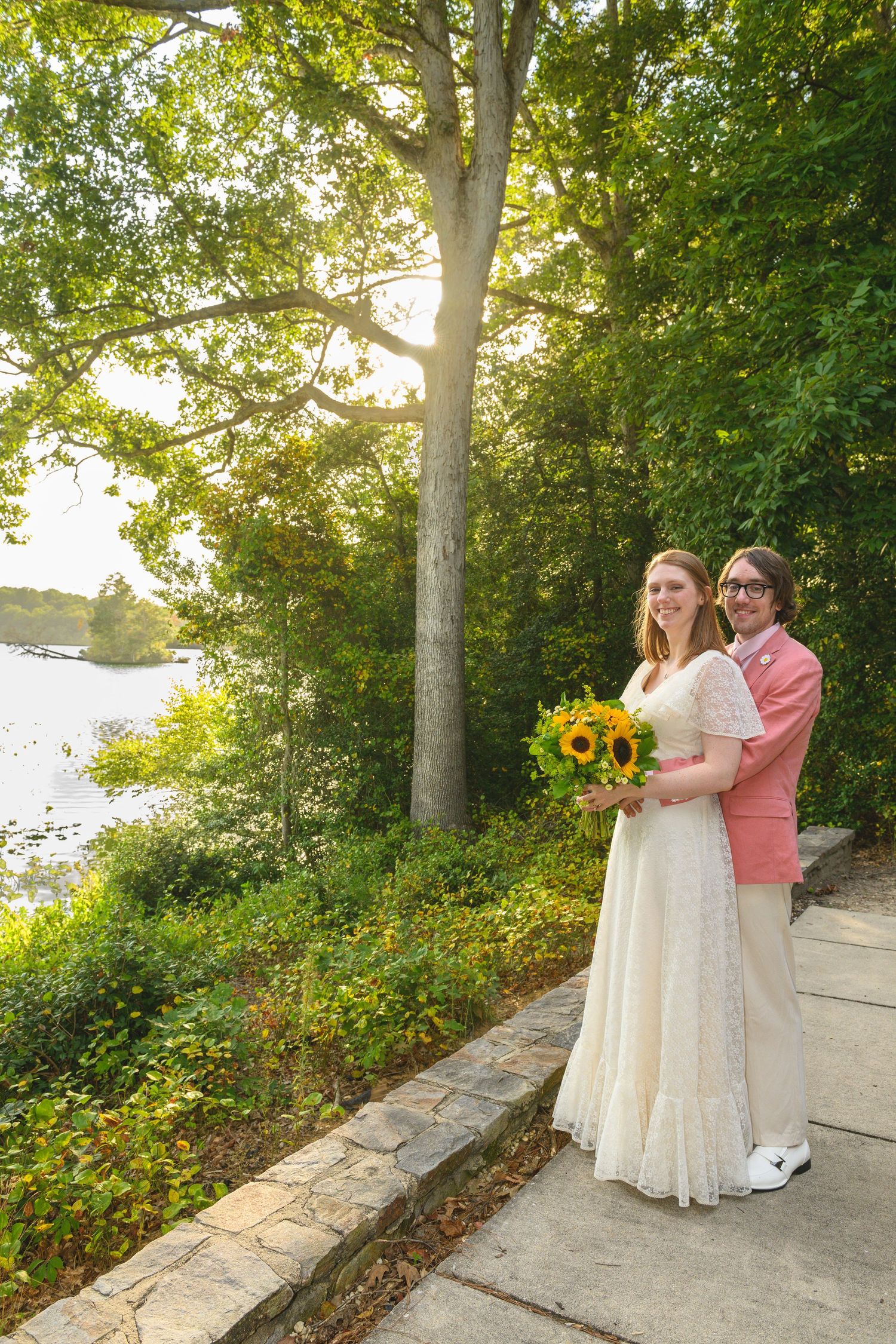 Bride and groom pose on stone path by sunlit lake, she holds sunflower bouquet, he wears pink jacket.