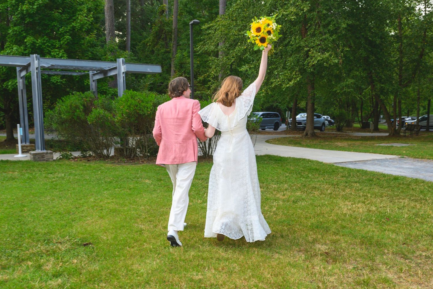Bride in white dress holding sunflowers walks with groom in pink suit across green lawn at outdoor wedding.