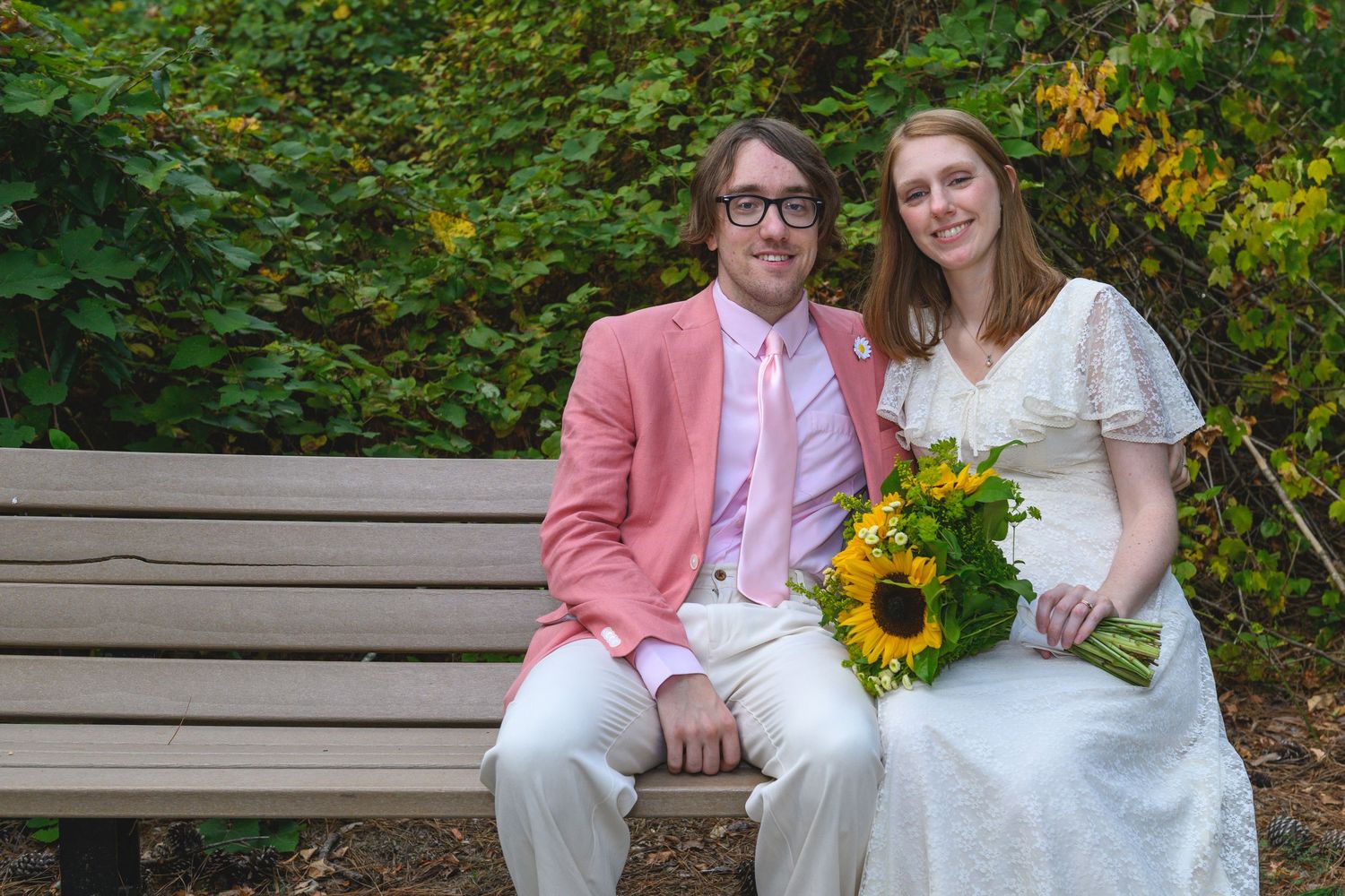 Couple in wedding attire sitting on bench, groom in pink suit, bride holding sunflower bouquet, green foliage background.