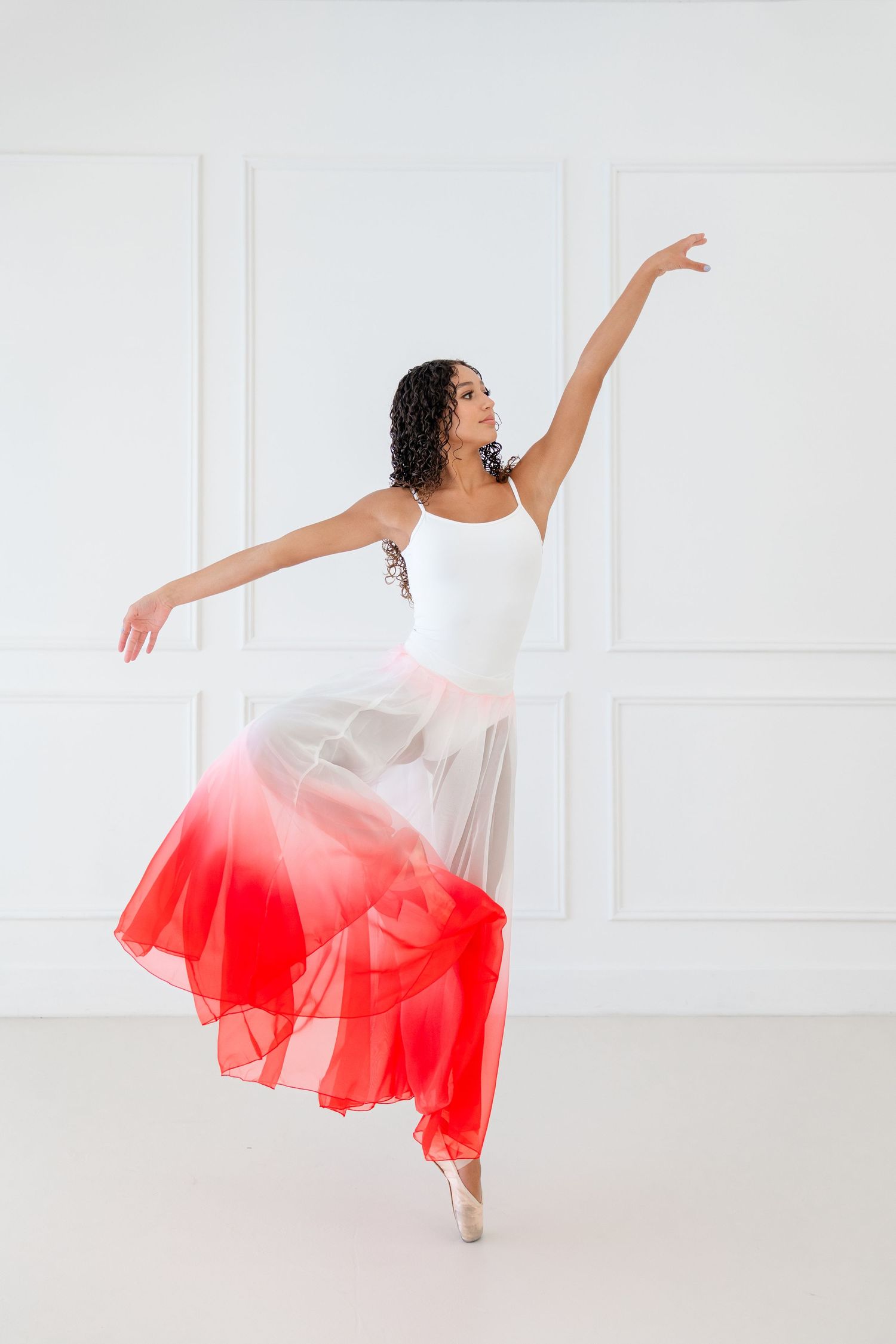 Ballerina in white leotard and red ombre skirt poses gracefully on pointe in a bright white studio.