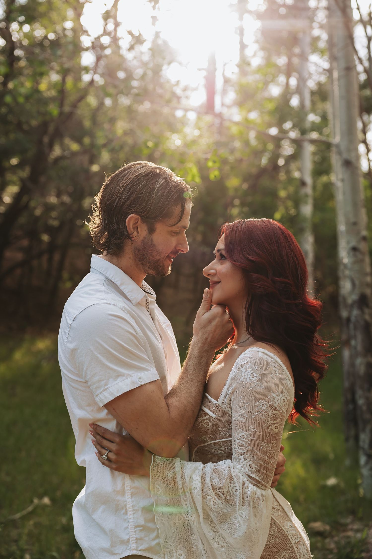 Couple sharing a romantic moment in a sunlit forest, woman in lace dress with red hair, man in white shirt.