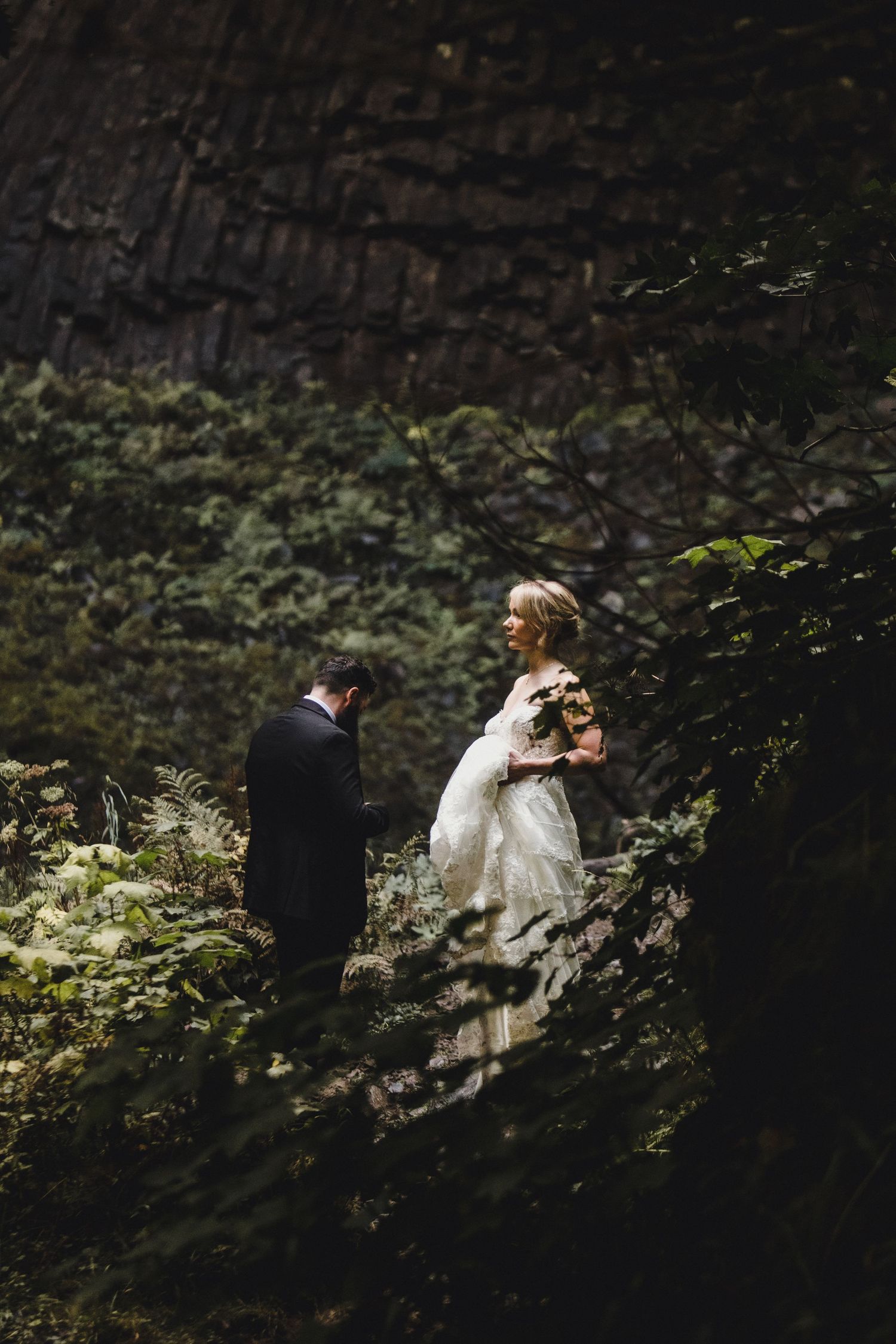 Bride and groom share intimate moment on rocky ledge in the Columbia River Gorge, surrounded by lush green forest and dark stone cliffs.