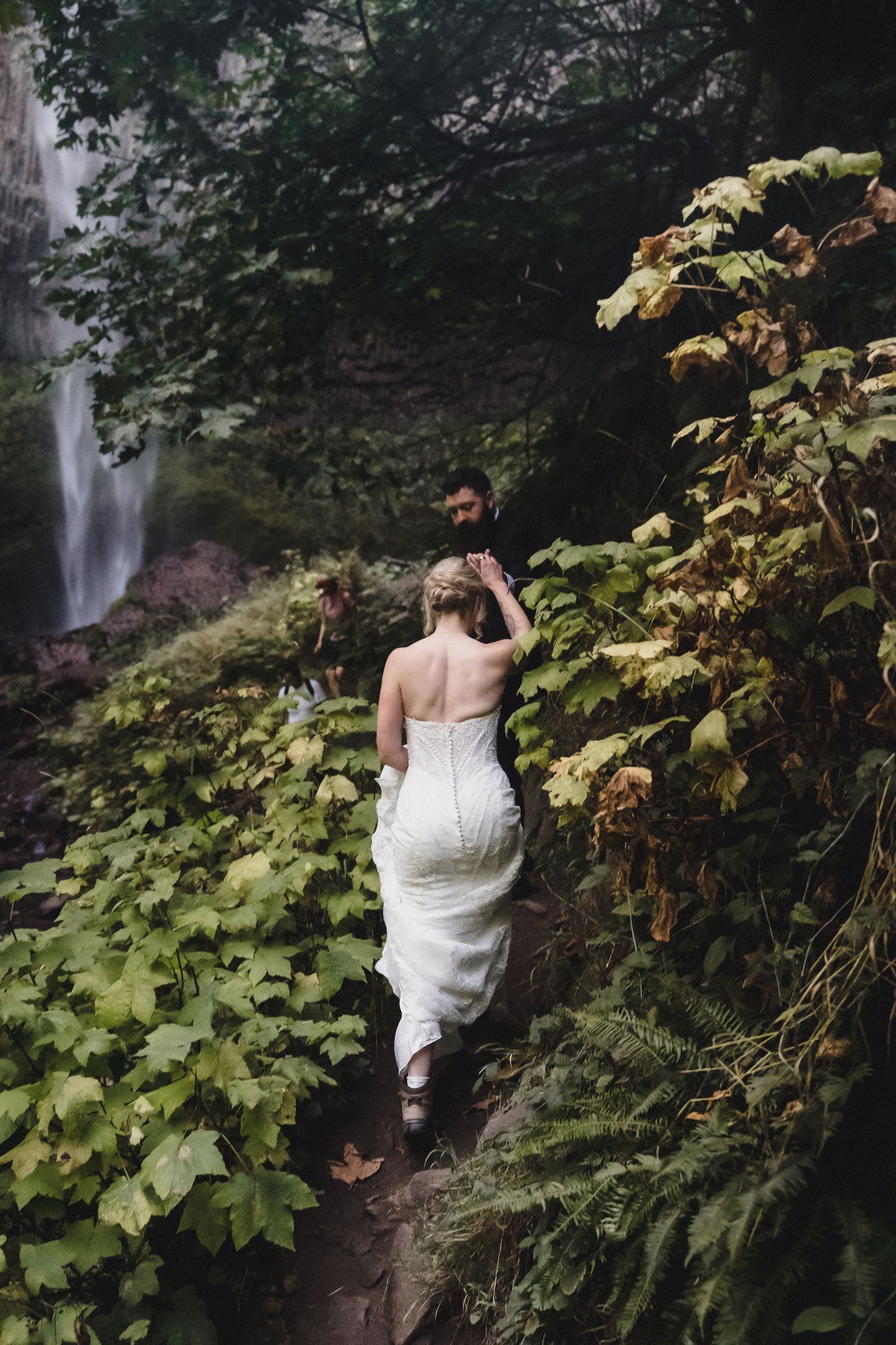 Bride in white gown and groom walk through lush forest path near waterfall during outdoor wedding elopement.
