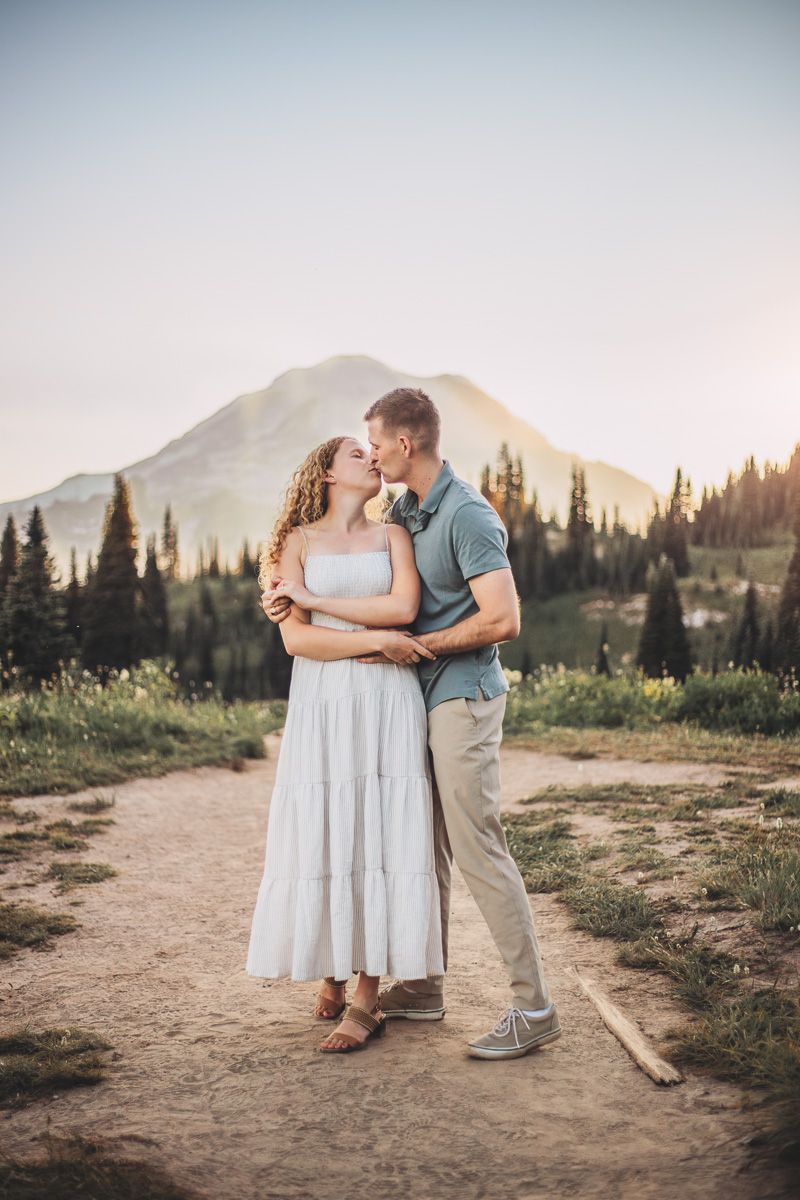 Couple sharing a romantic kiss on a Mt. Rainier mountain trail with evergreen trees and a scenic peak at golden hour.